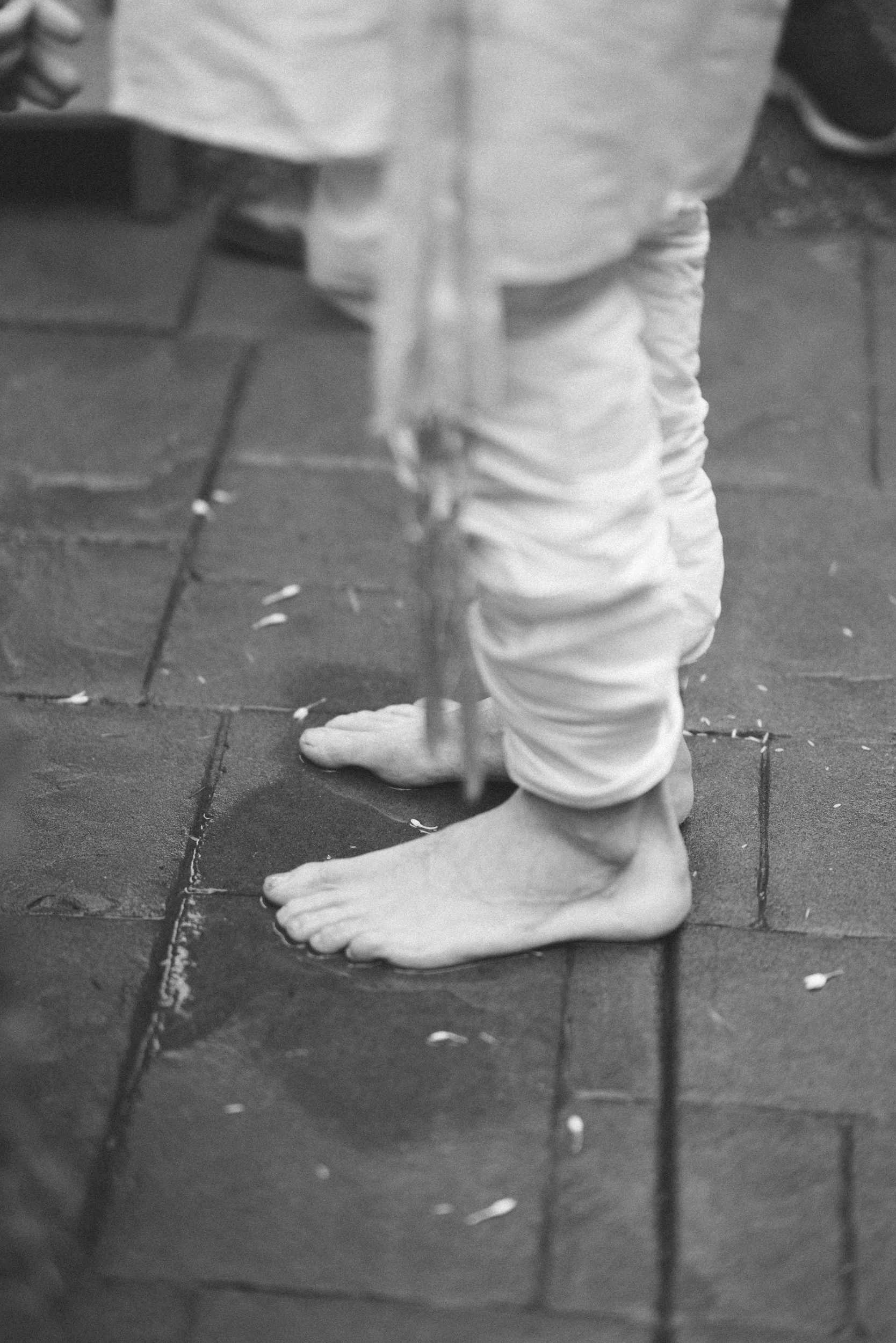 Close-up of a person barefoot on tile flooring, wearing light-colored pants, with part of other person's clothes and shoes visible in the background.