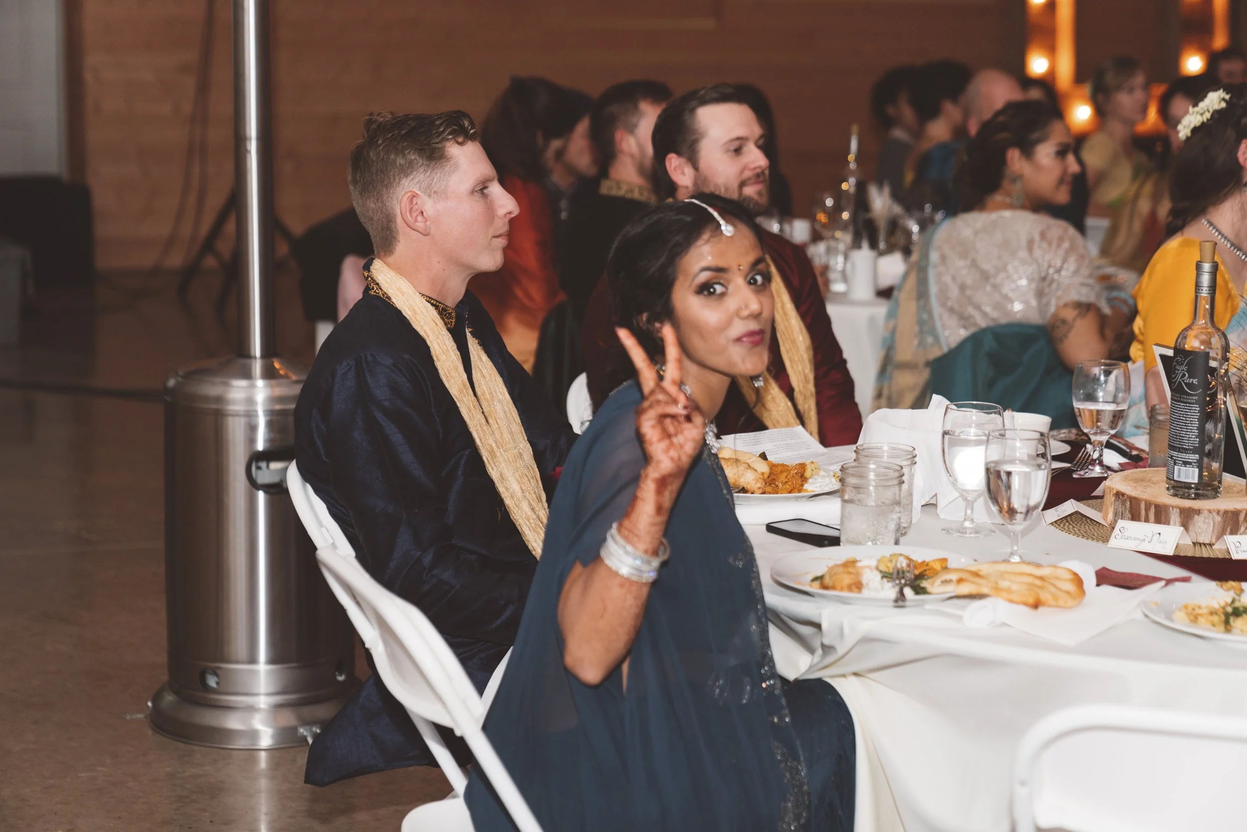 People seated at a decorated dining table during a celebration, including a woman making a peace sign and smiling at the camera.