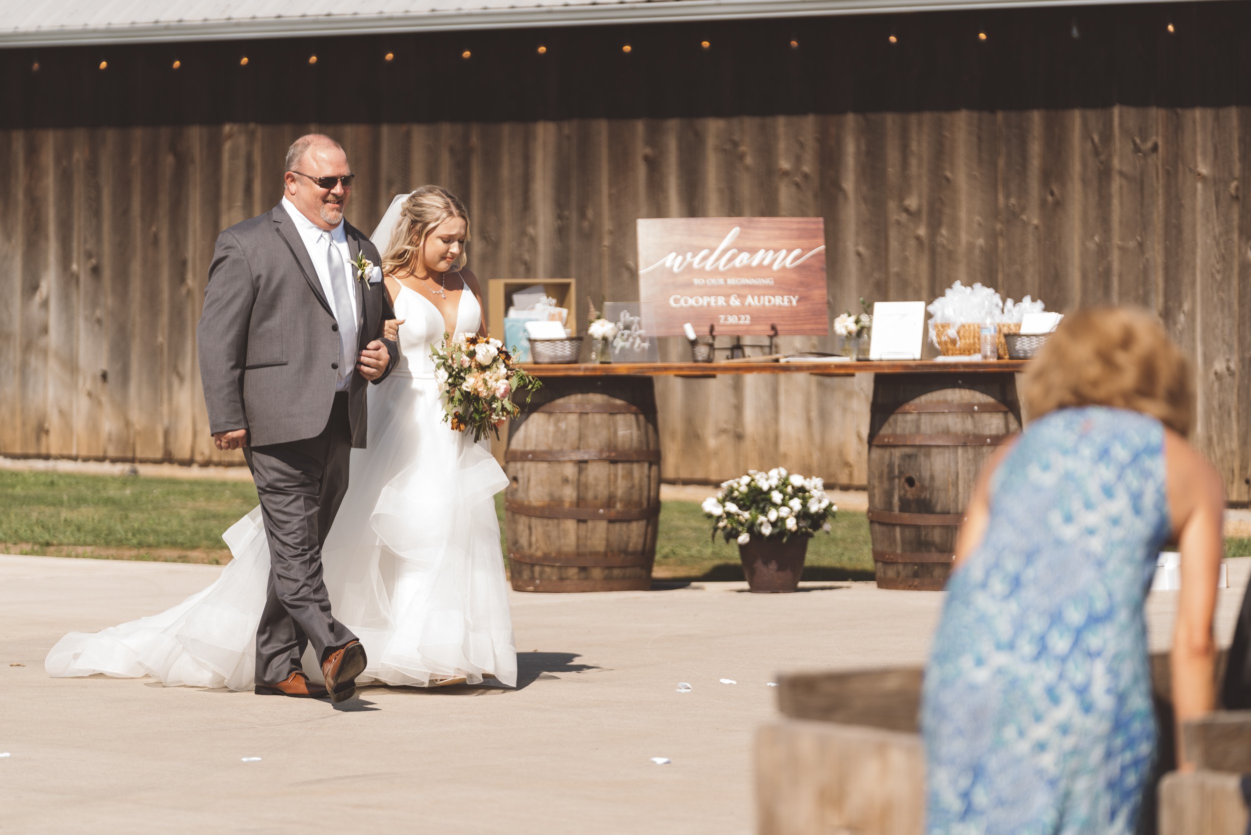 A bride walking down the aisle with a man holding her arm at a wedding ceremony, with a wooden backdrop and a table decorated with flowers and a welcome sign for Cooper and Audrey.