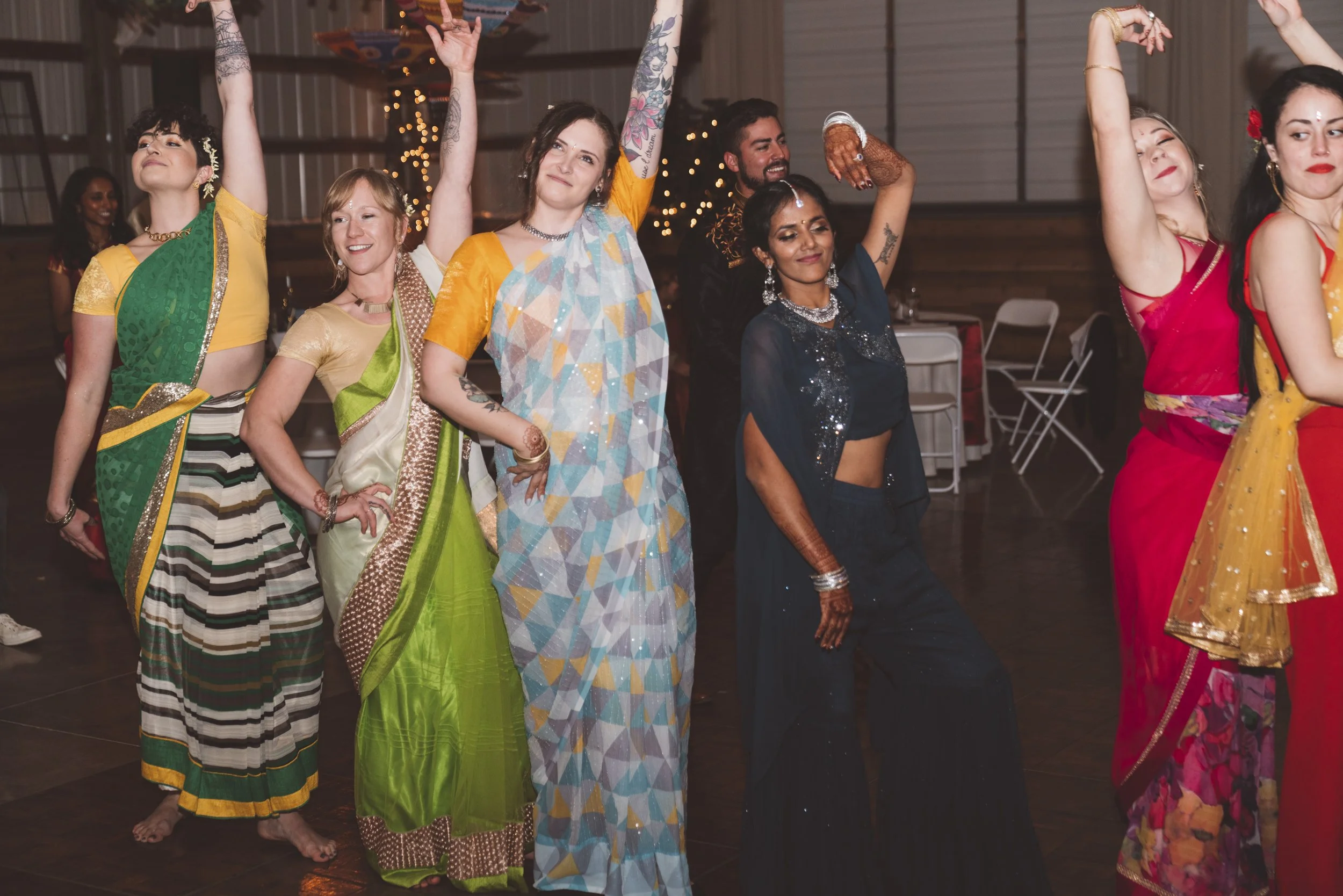Group of seven women dancing together at an event, dressed in colorful traditional Indian sarees and salwar kameez, standing in a line, smiling and raising their arms, with festive lights in the background.