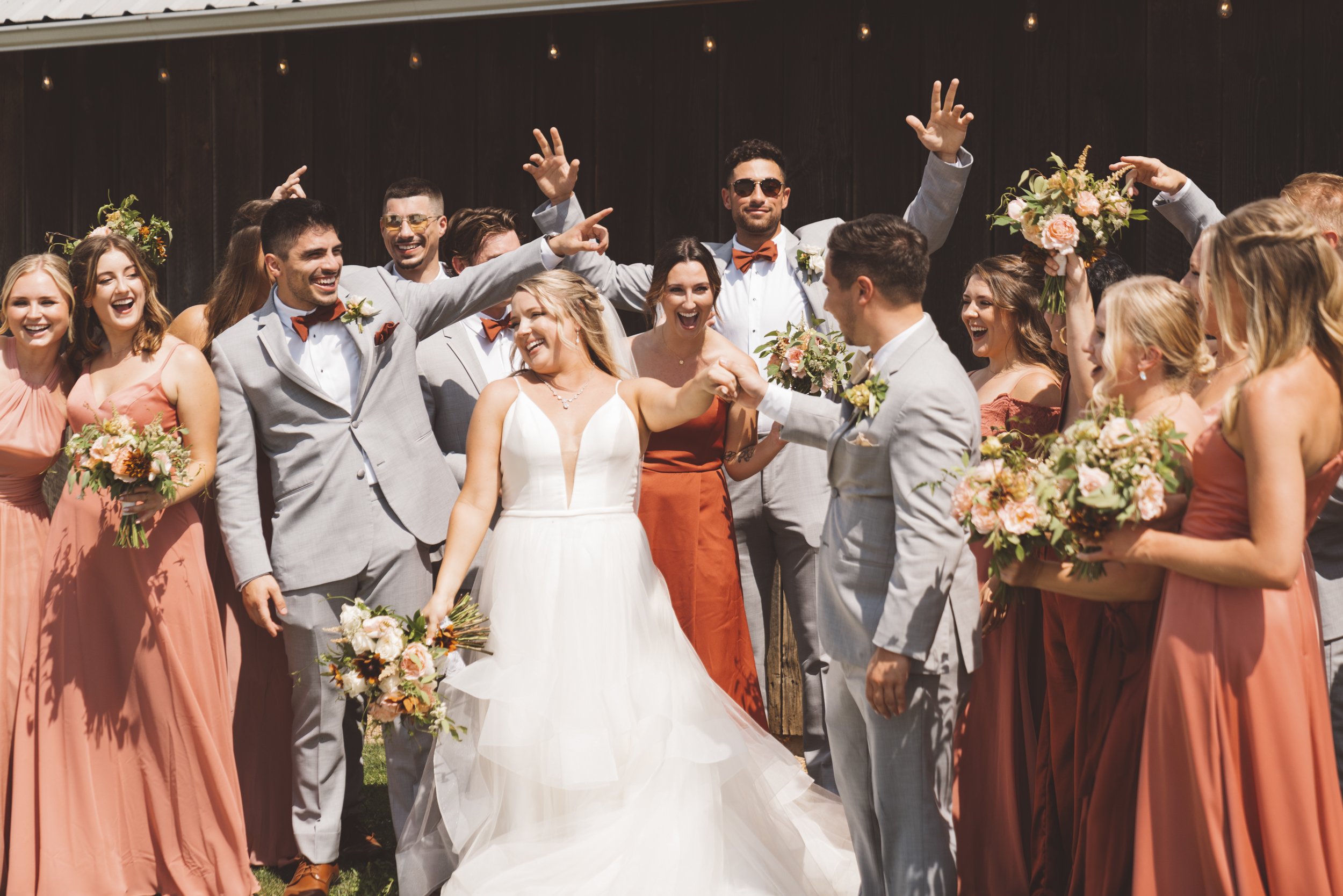 A wedding party celebrating outdoors, with the bride and groom in the center surrounded by bridesmaids and groomsmen, all smiling and holding bouquets of flowers.