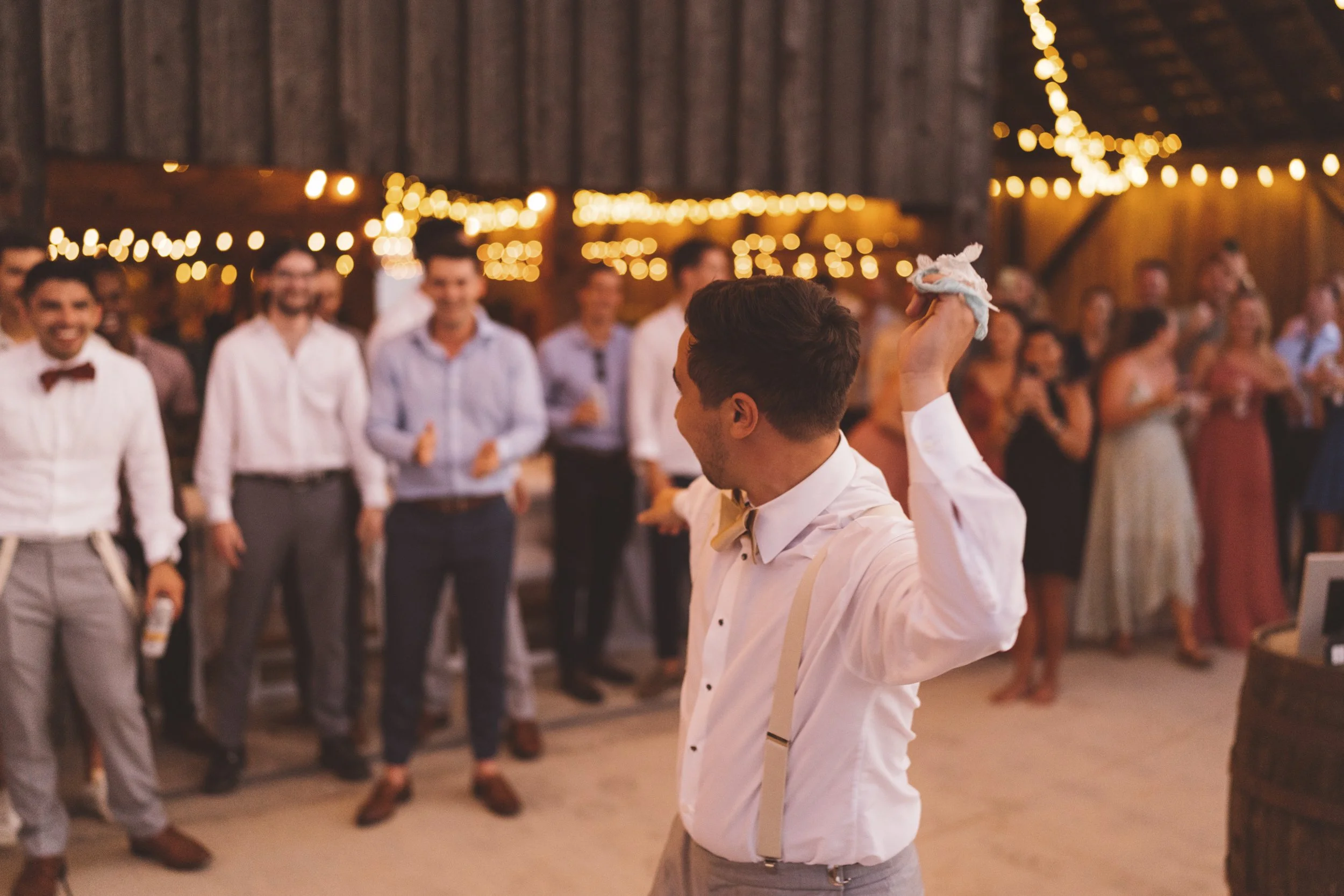 A man in a white shirt and suspenders holding a sock while dancing at a party or wedding reception, with a crowd of people in the background watching and smiling under string lights in a barn-like venue.