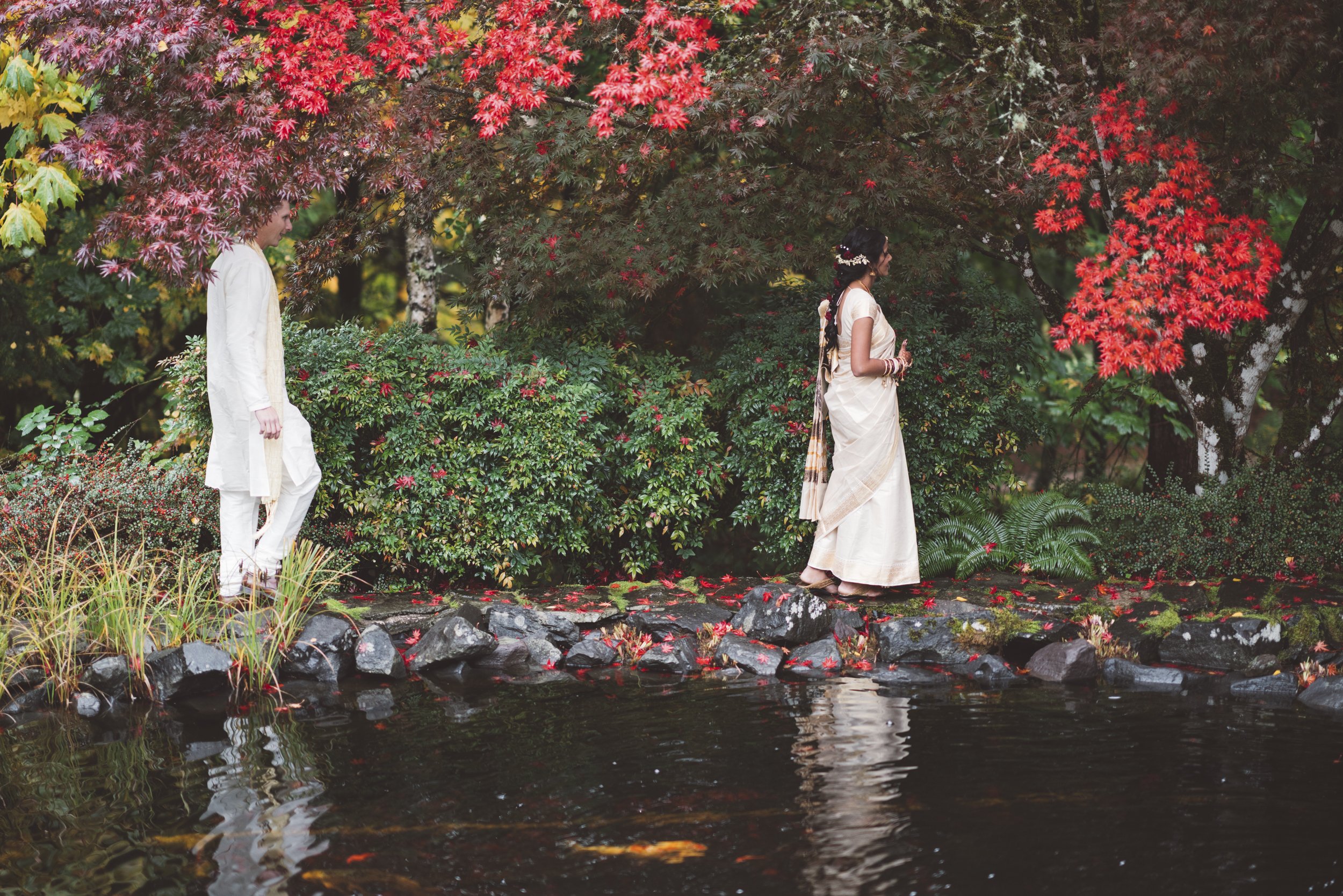 A man and woman dressed in traditional Indian attire walking along a rocky edge next to a pond, surrounded by vibrant autumn foliage.
