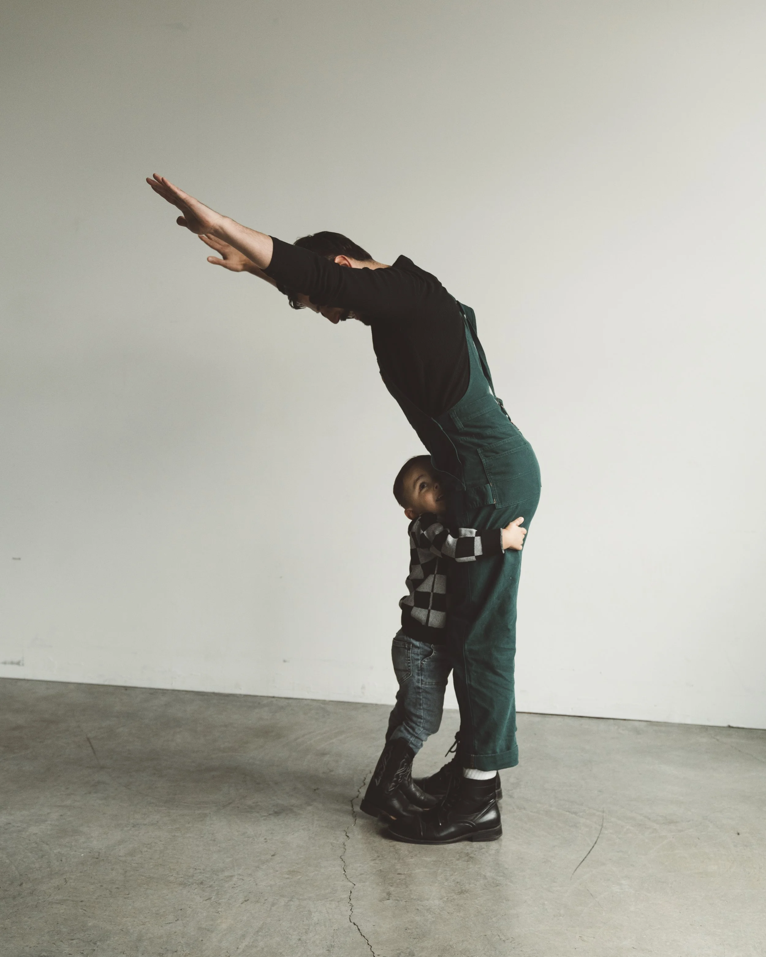 A woman standing on her tiptoes with her arms extended forward, while a young boy hugs her legs and looks up at her.
