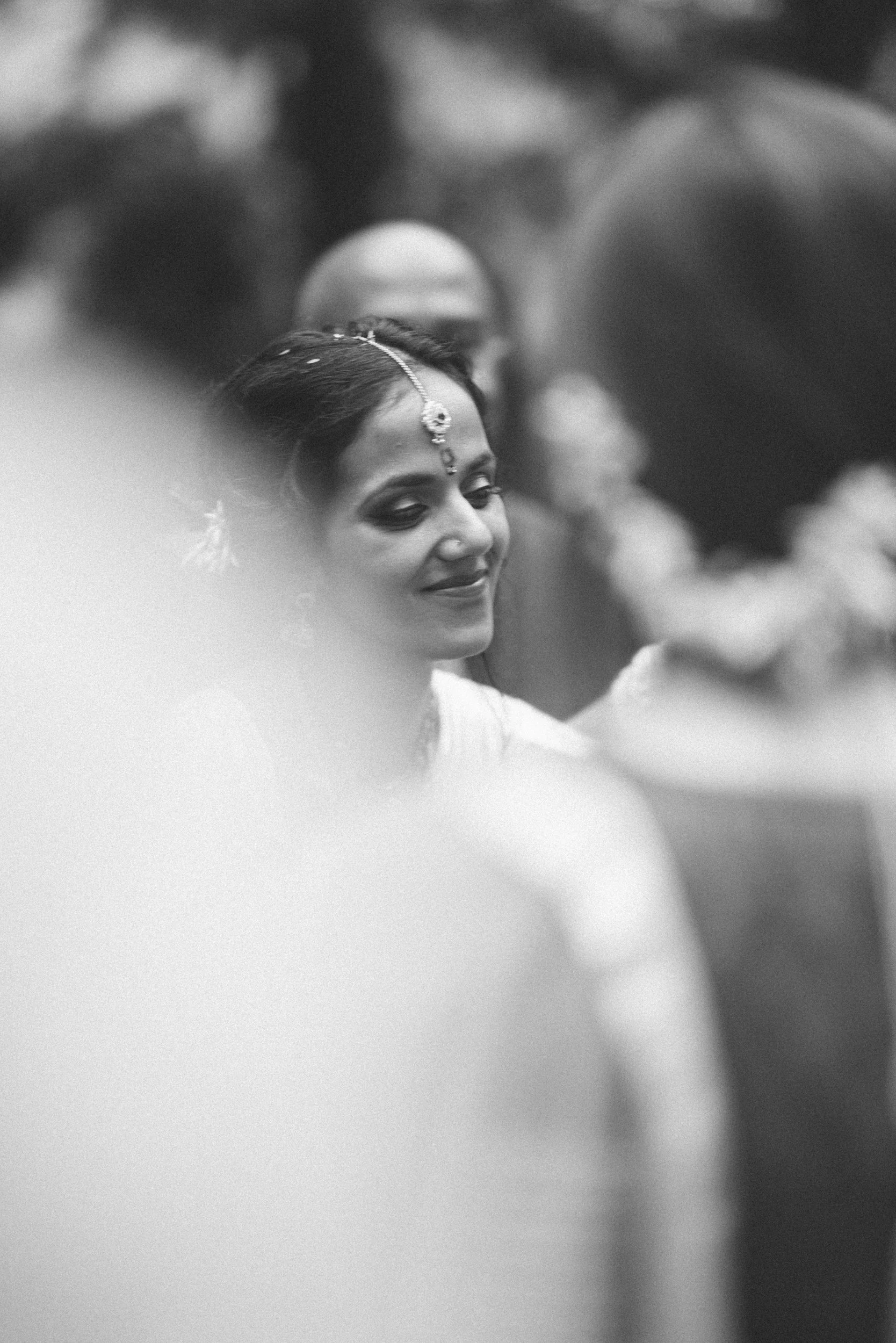 A woman dressed in traditional Indian attire, with jewelry and a bindi, smiling with her eyes closed amidst a crowd of people.