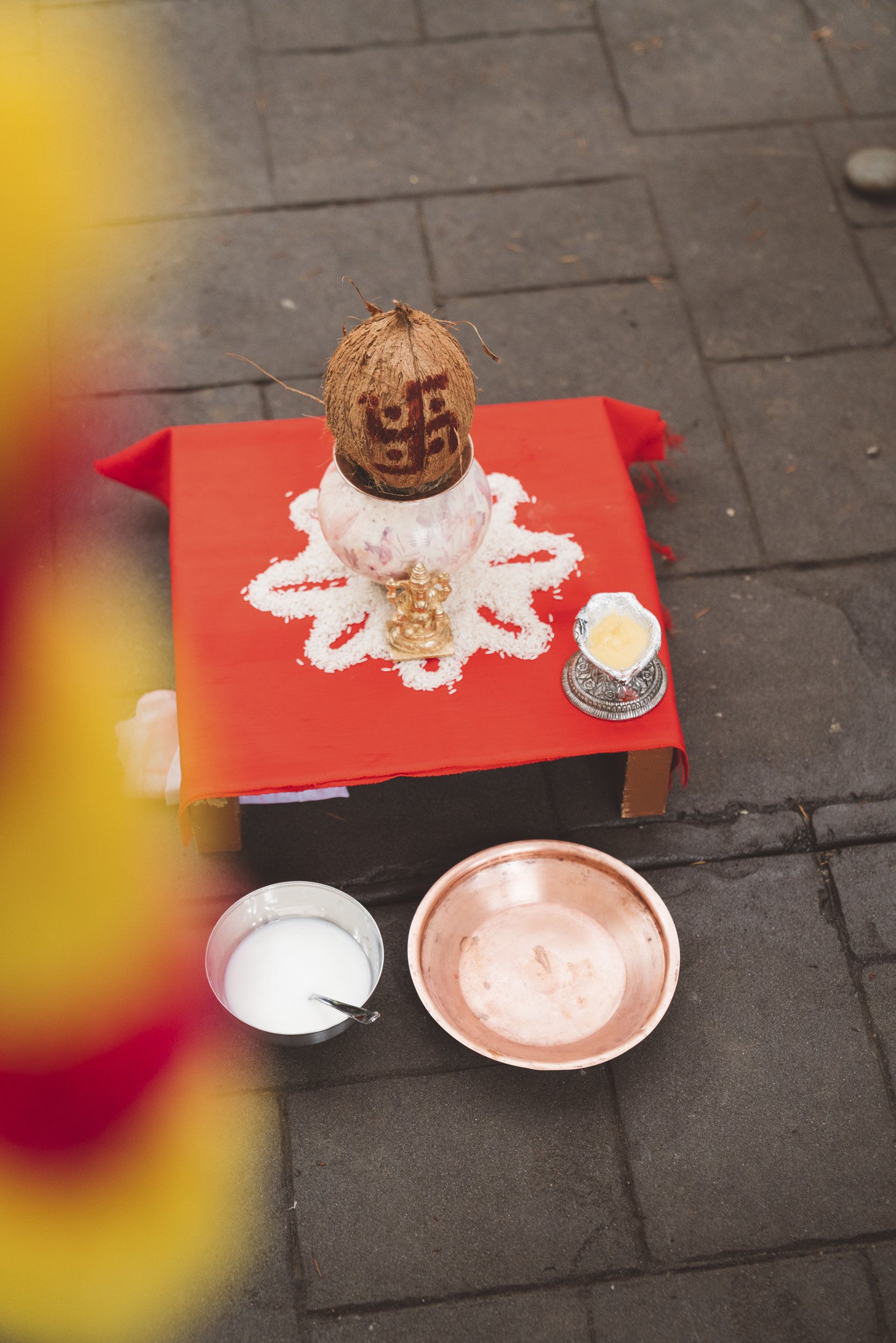 A small altar setup on a sidewalk with a coconut, a small statue, and bells, surrounded by bowls of milk and a small dish of butter, on a red cloth.