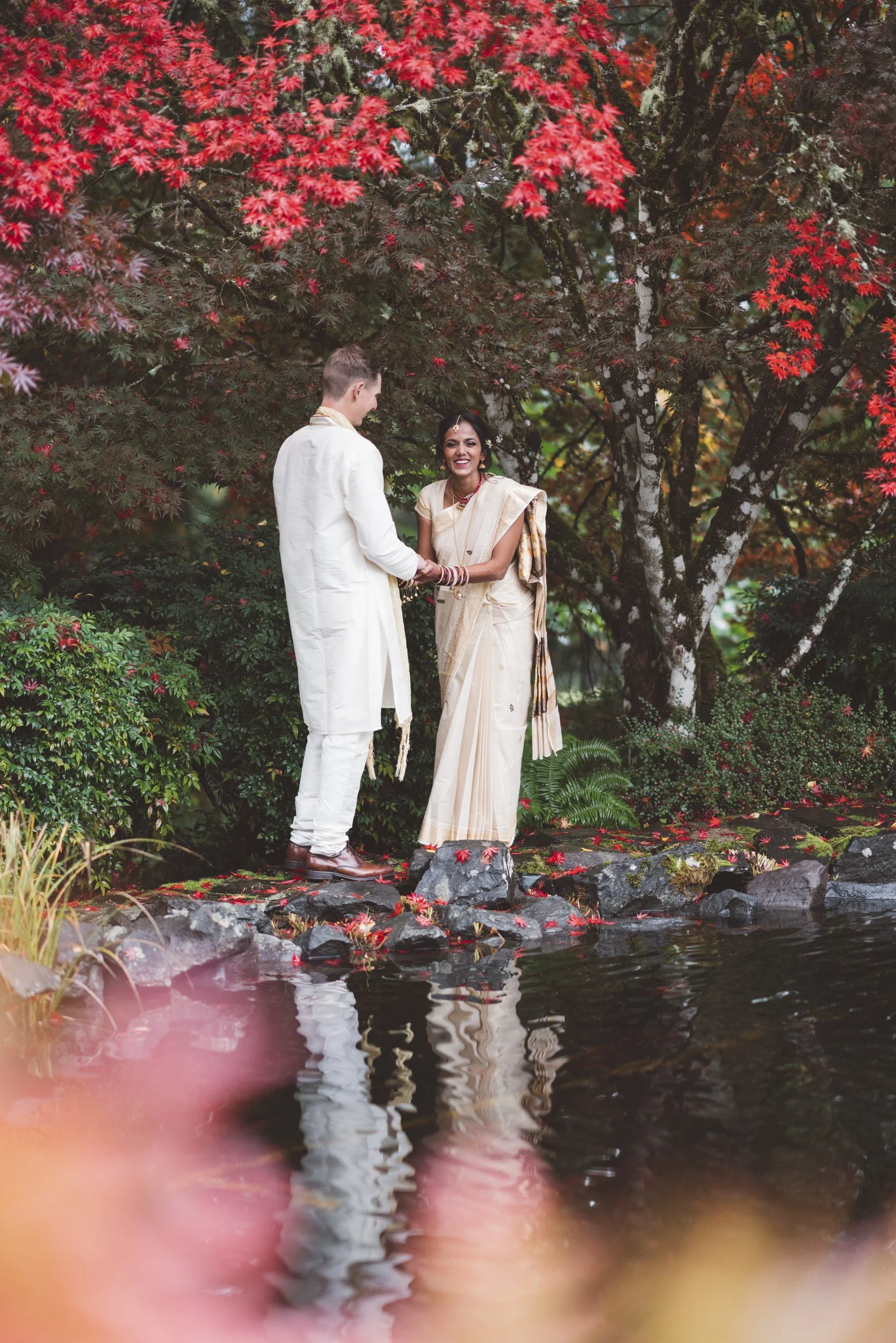 A couple dressed in traditional Indian attire stands on rocks by a pond, exchanging rings, with vibrant red and green trees in the background.
