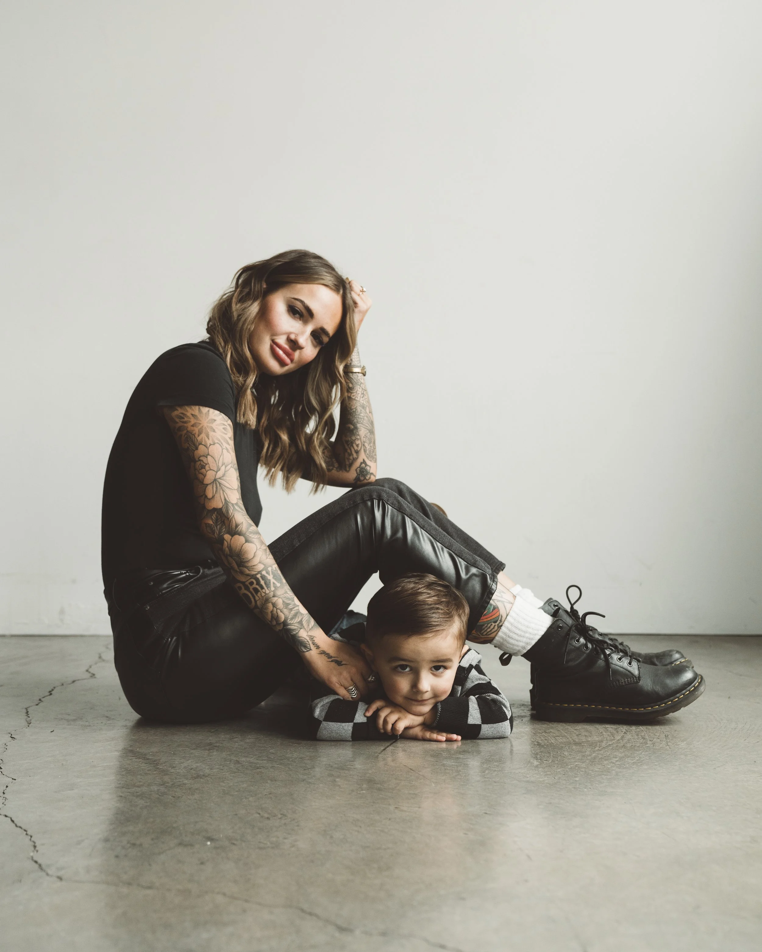 A woman with tattoos and wearing black leather pants and boots, sitting on the floor with her young son, who is lying on his stomach with his head resting on his hands. They are in a plain room with a concrete floor and white wall.
