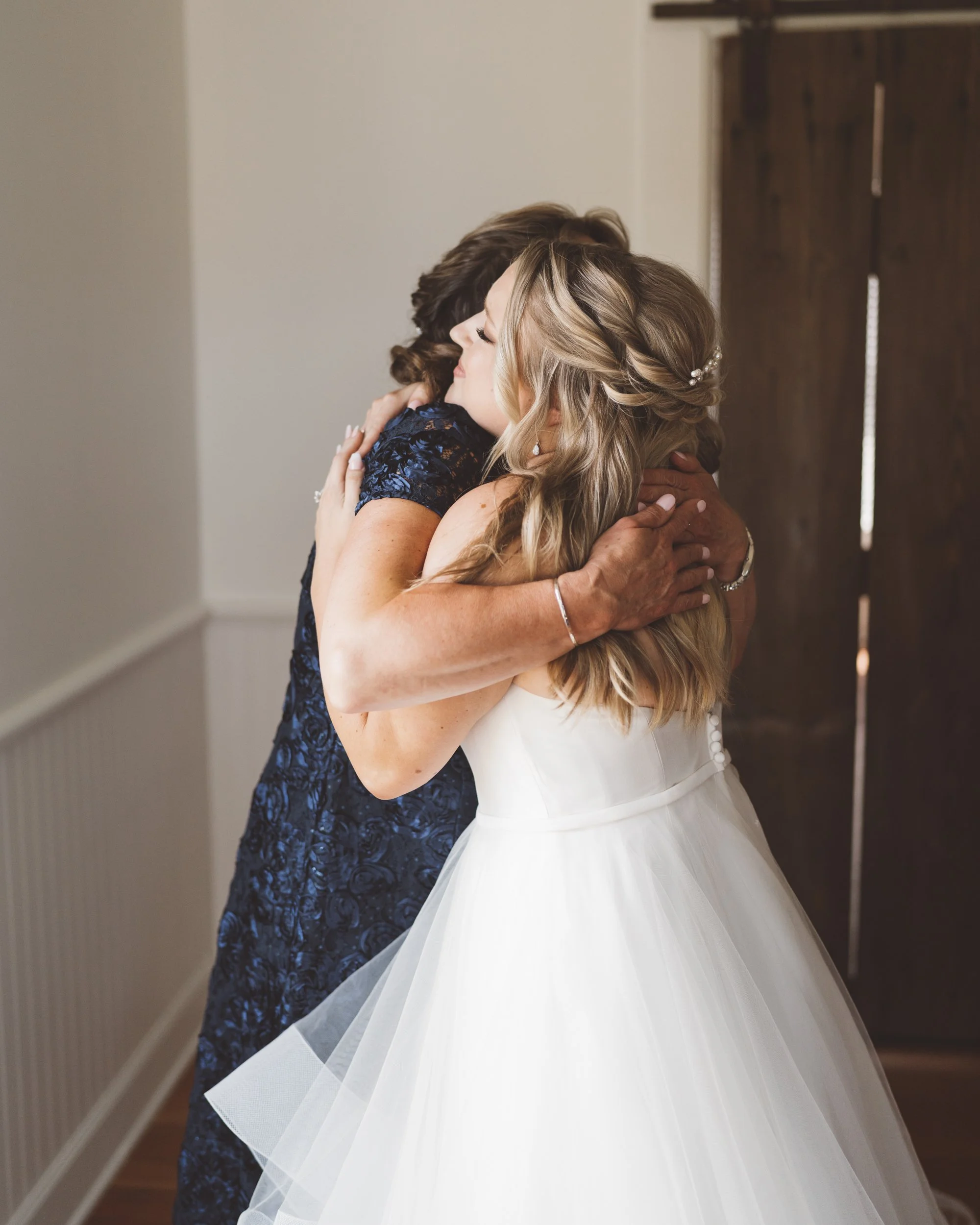 A woman in a white wedding dress hugging another woman, possibly her mother, in a blue dress, inside a room with wooden door and white walls.
