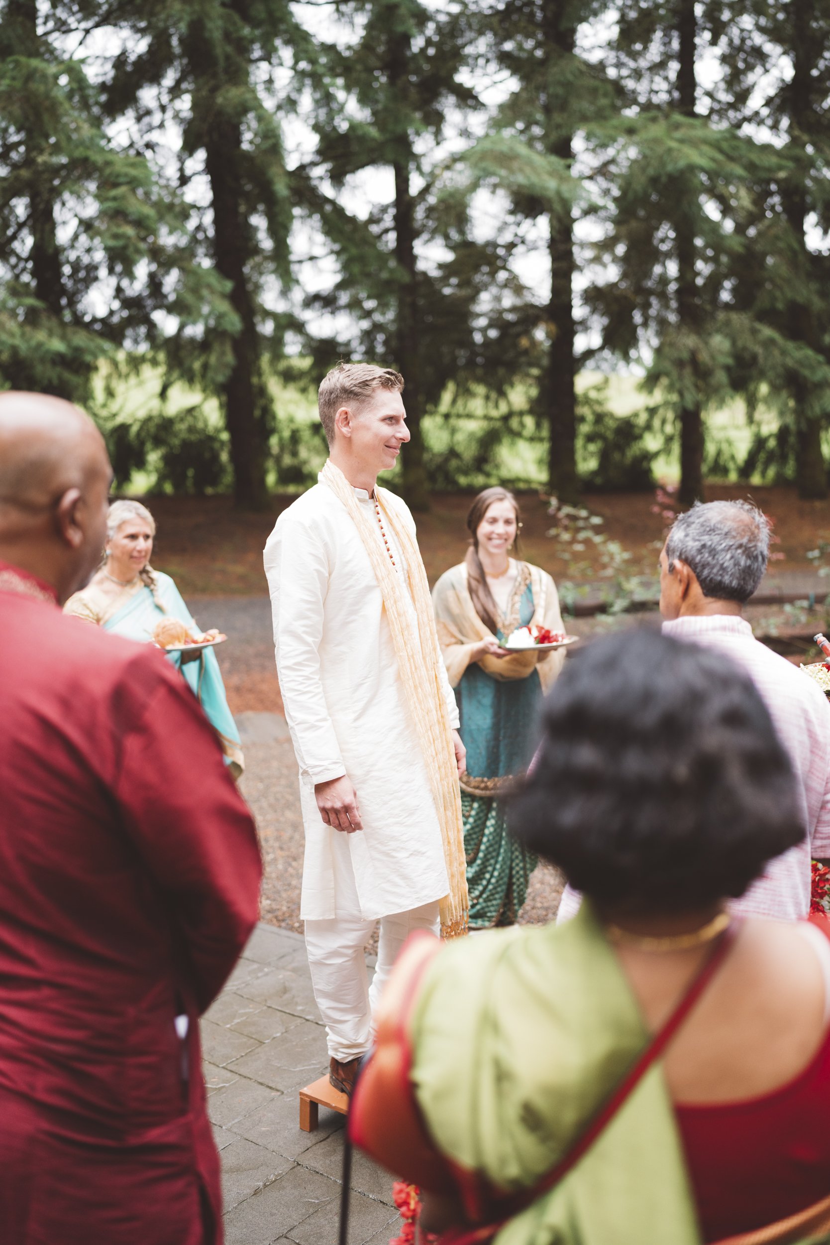 A group of people participating in a traditional Hindu wedding ceremony outdoors, with the groom standing on a small wooden platform, dressed in traditional cream-colored attire, while others hold offerings, surrounded by lush green trees.