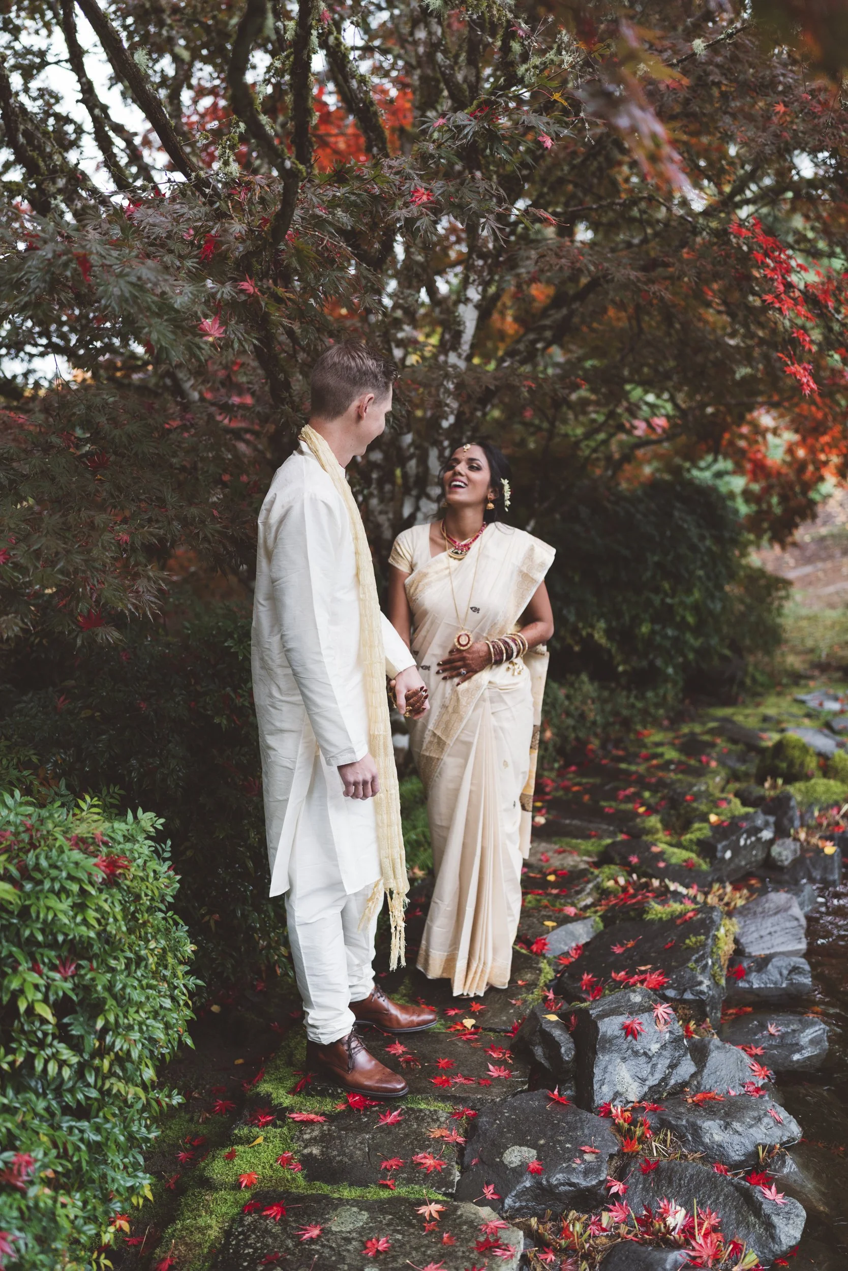 A couple dressed in traditional Indian attire standing on a stone pathway surrounded by autumn-colored trees and fallen leaves, holding hands and smiling.