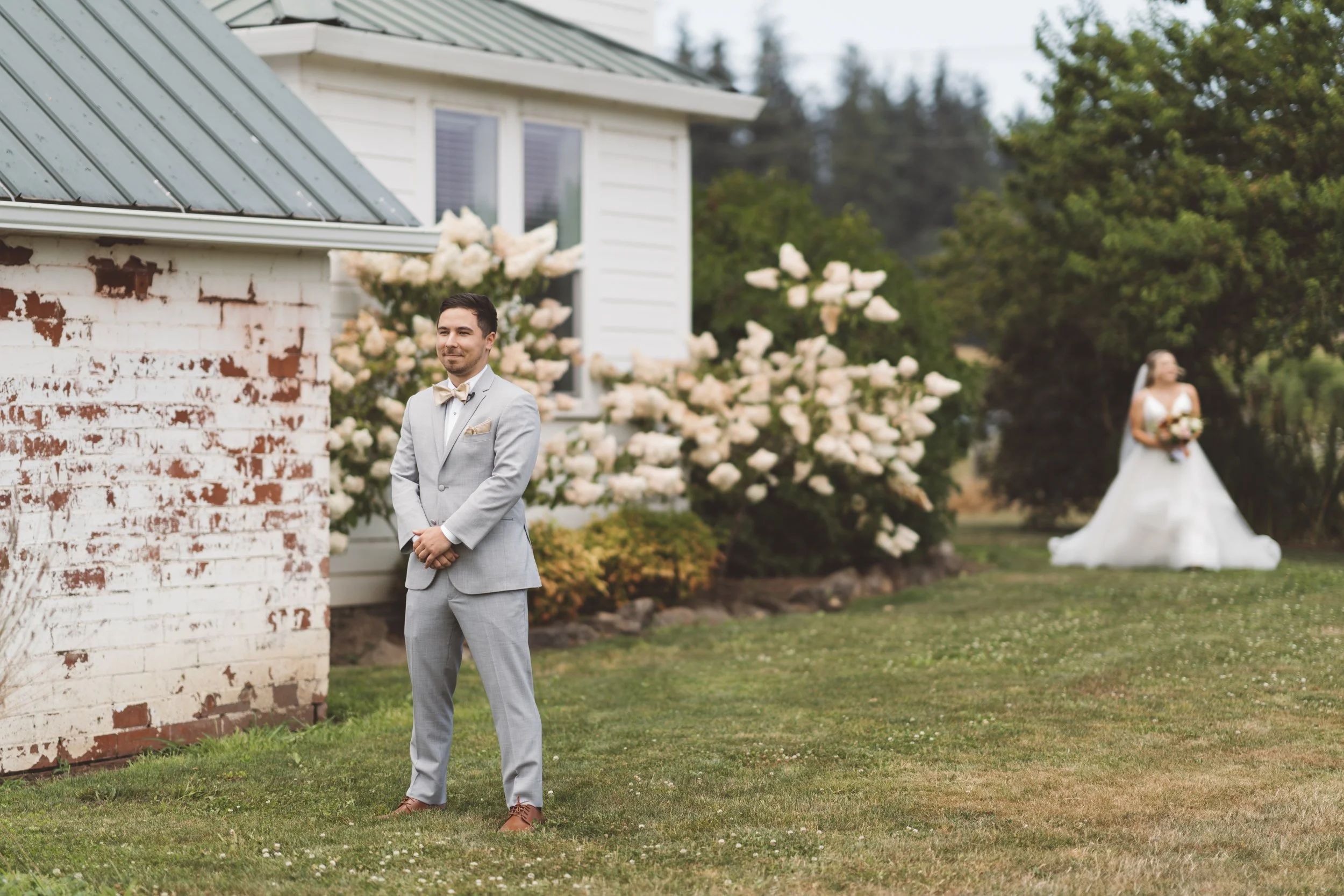 A groom in a light gray suit standing outdoors, with a bride in a white wedding dress in the background near bushes with white flowers.