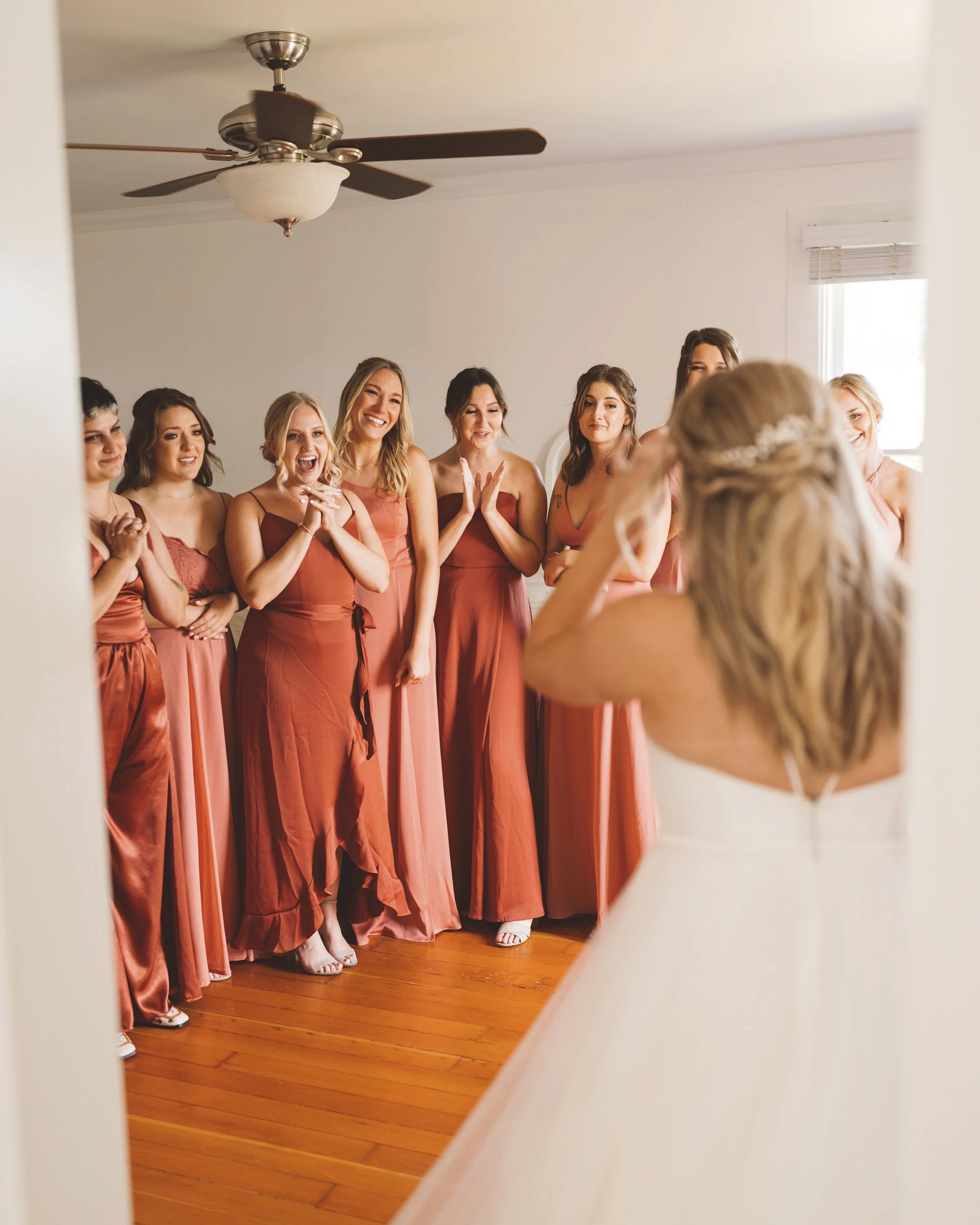 A bride in a white wedding dress is looking at her bridesmaids who are all dressed in matching rust-colored dresses, celebrating and smiling in a room with wooden floors and a ceiling fan.