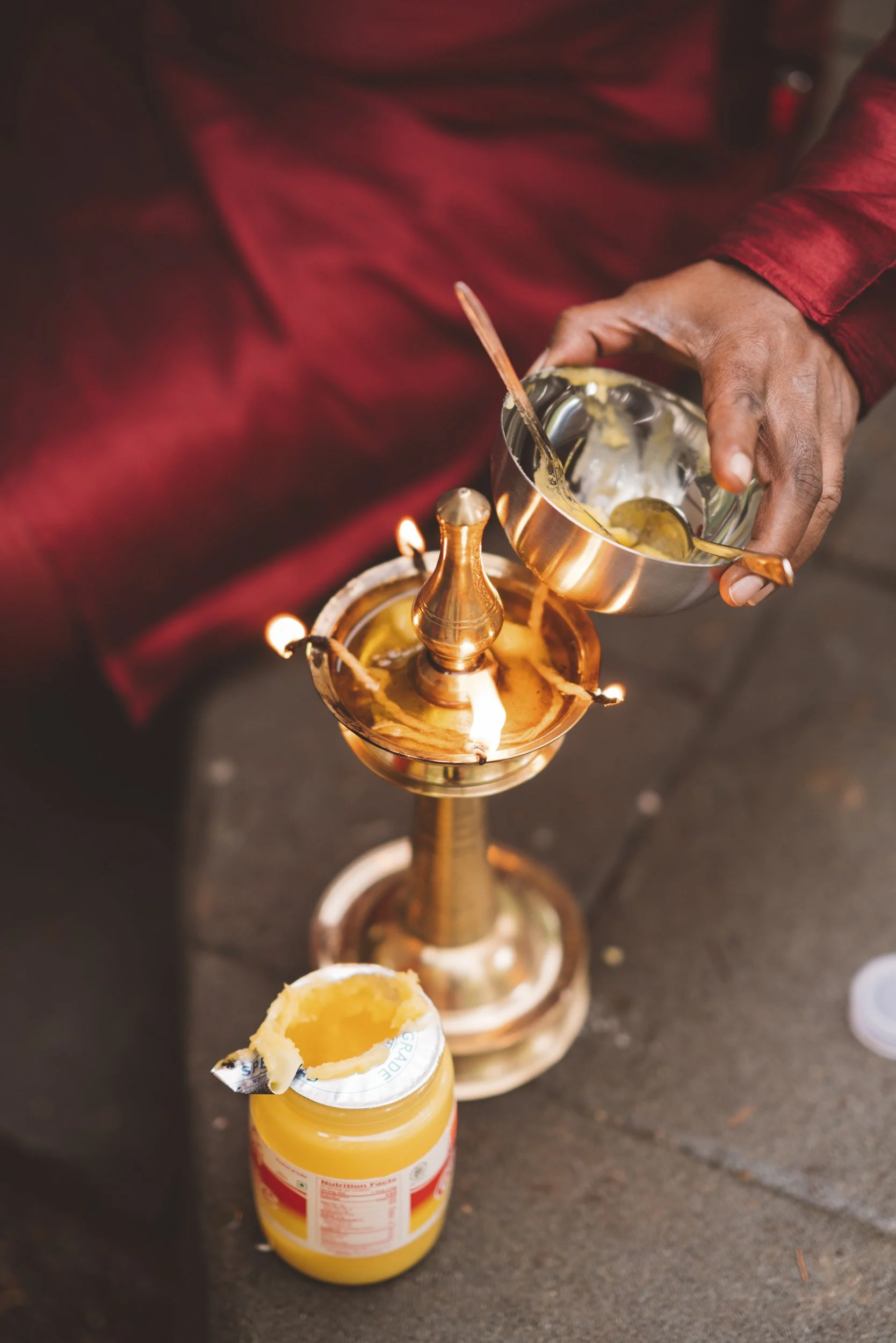 Person pouring butter from a metal bowl into a lit brass oil lamp with a small pot of yellow substance nearby.