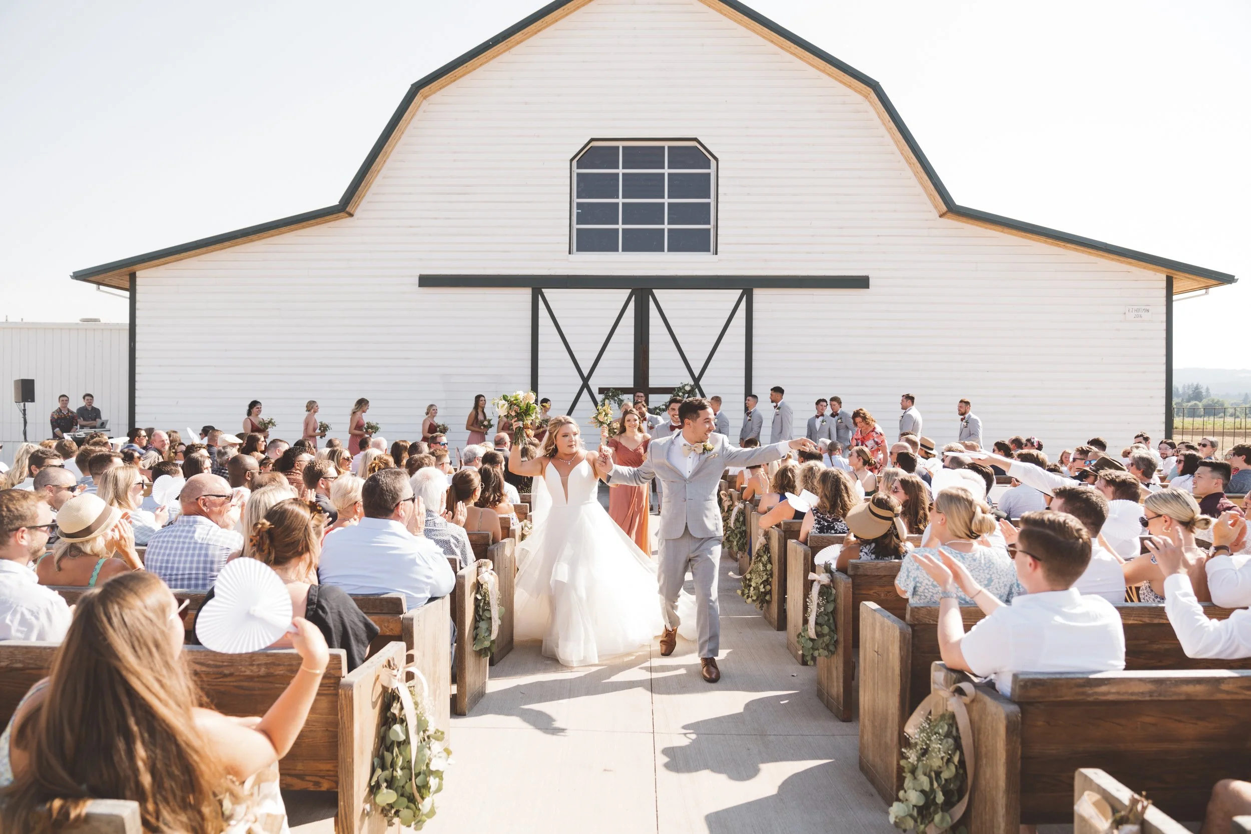 Outdoor wedding ceremony in front of a large white barn with a black sliding door, where a bride and groom are walking down the aisle, surrounded by seated guests.