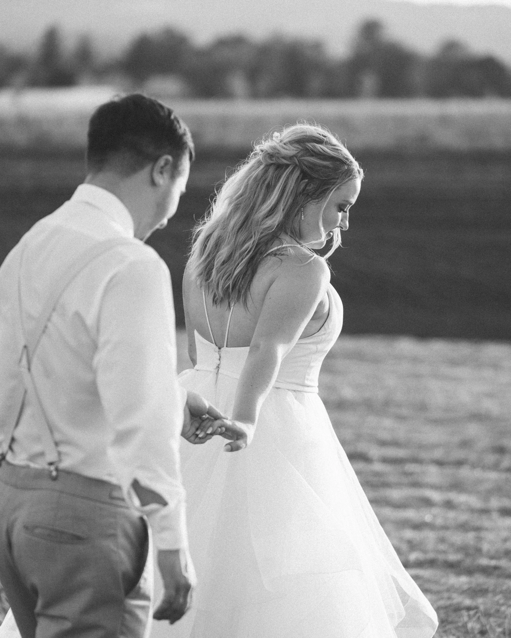 A black and white photo of a man and woman holding hands outdoors, with the woman in a wedding dress and the man in suspenders and a white shirt.