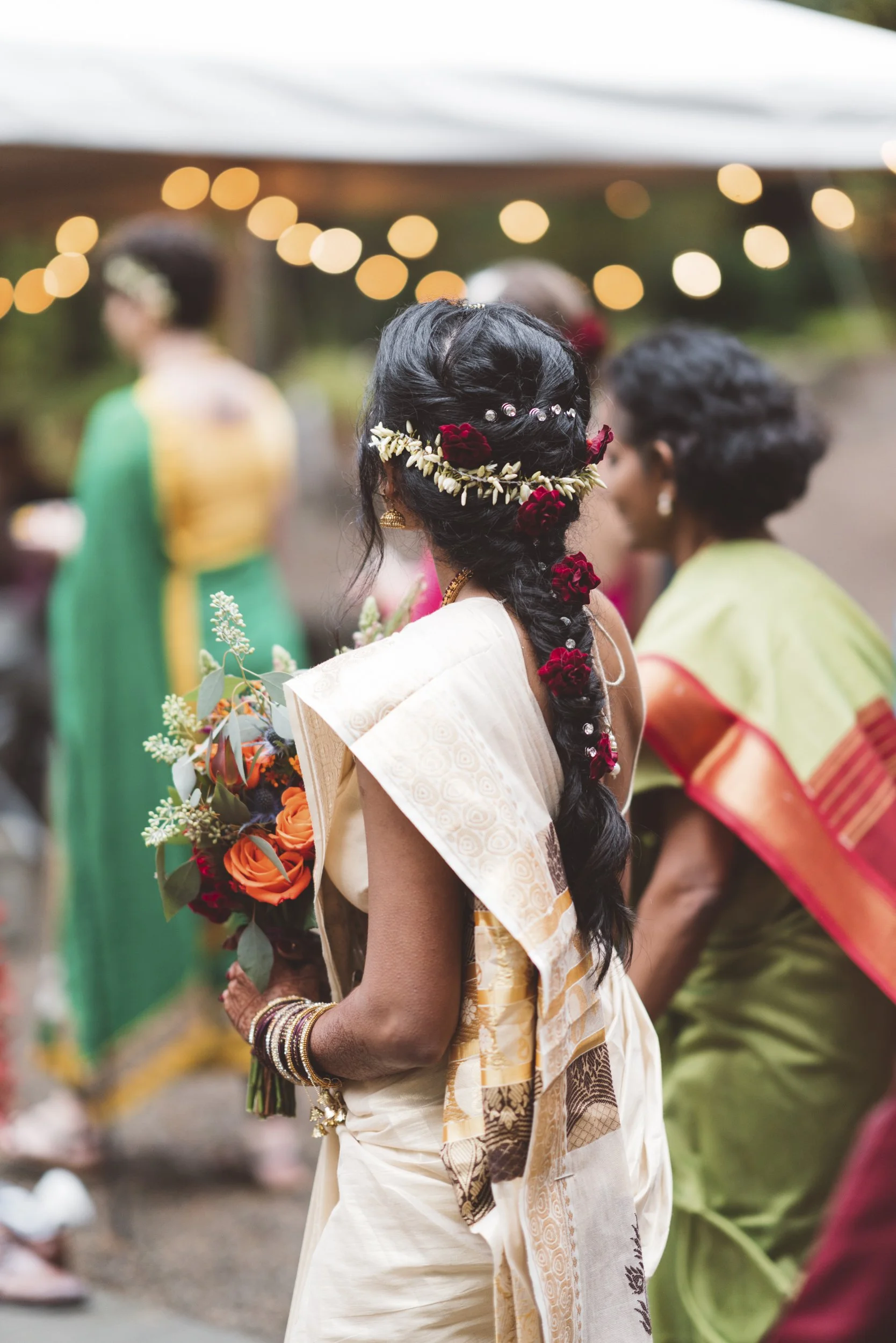 A woman in traditional Indian attire with floral jewelry and a braid decorated with flowers, holding a bouquet of flowers at a celebration outdoors, with blurred guests and string lights in the background.