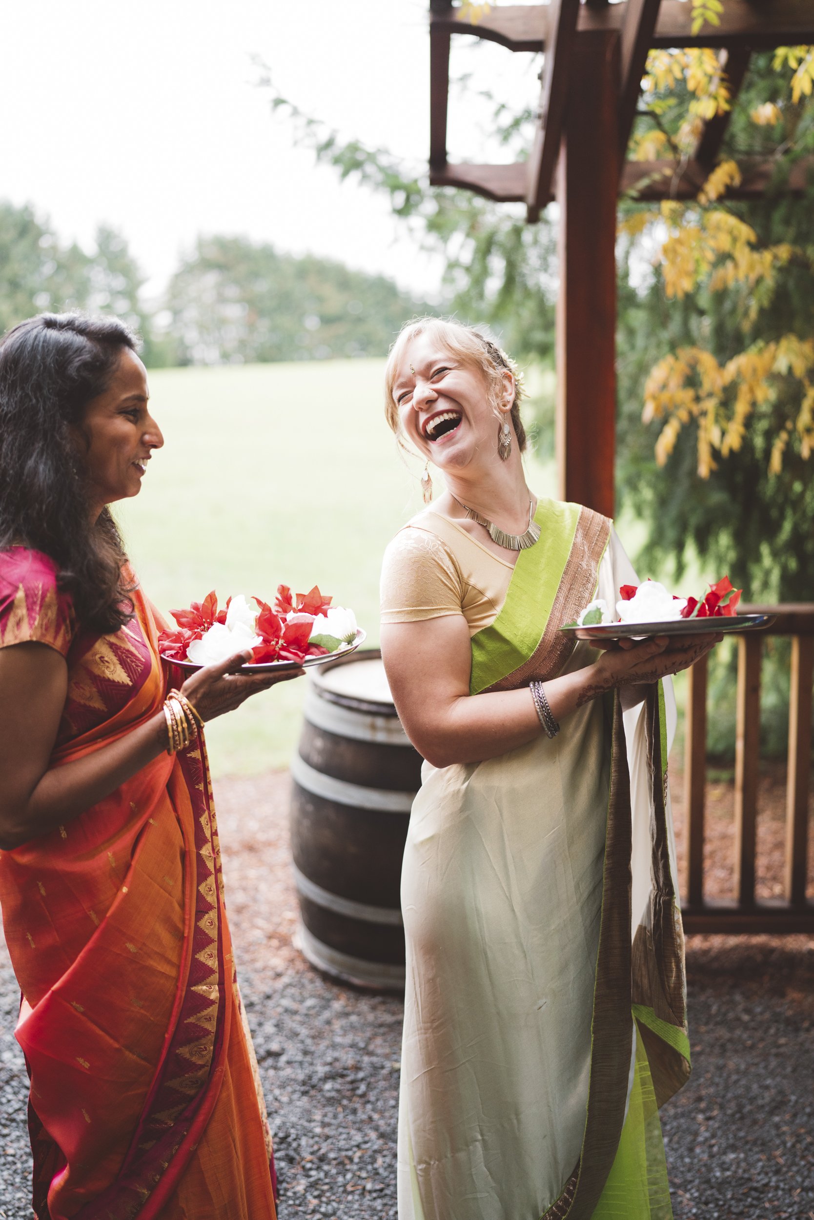 Two women dressed in traditional Indian sarees sharing a joyful moment while holding plates of desserts outdoors, with a wooden structure and green landscape in the background.