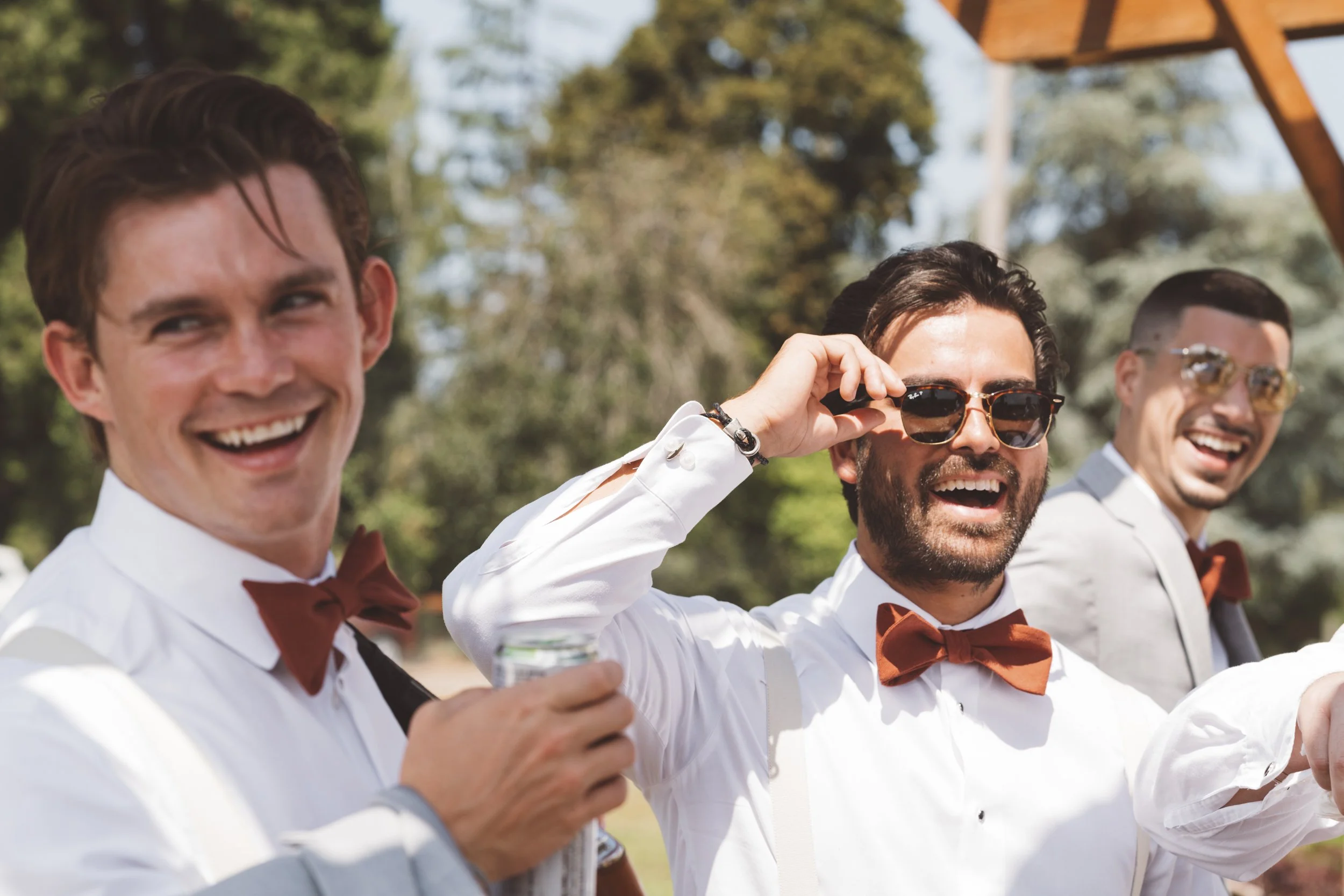 Three men dressed in white shirts with brown bow ties enjoying outdoors, one is holding a drink, and another is adjusting sunglasses, with trees and sky in the background.