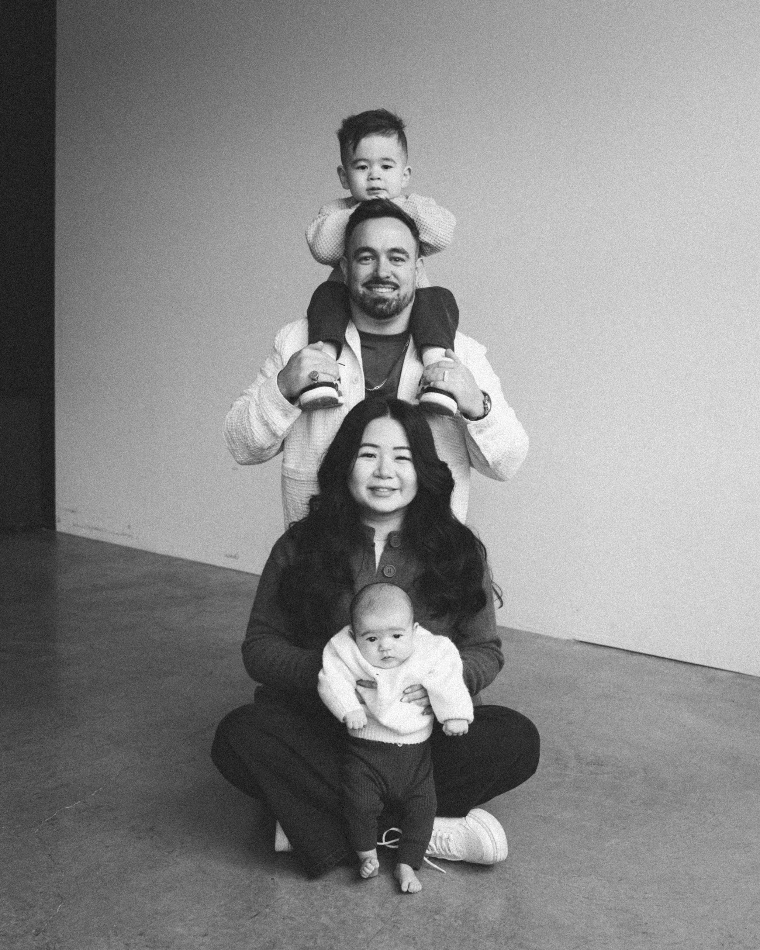 Black and white photograph of a family with four members: a woman sitting cross-legged on the floor holding a baby, a man sitting behind her, one child sitting on the man's shoulders, and another child sitting on the woman's lap. They are all smiling