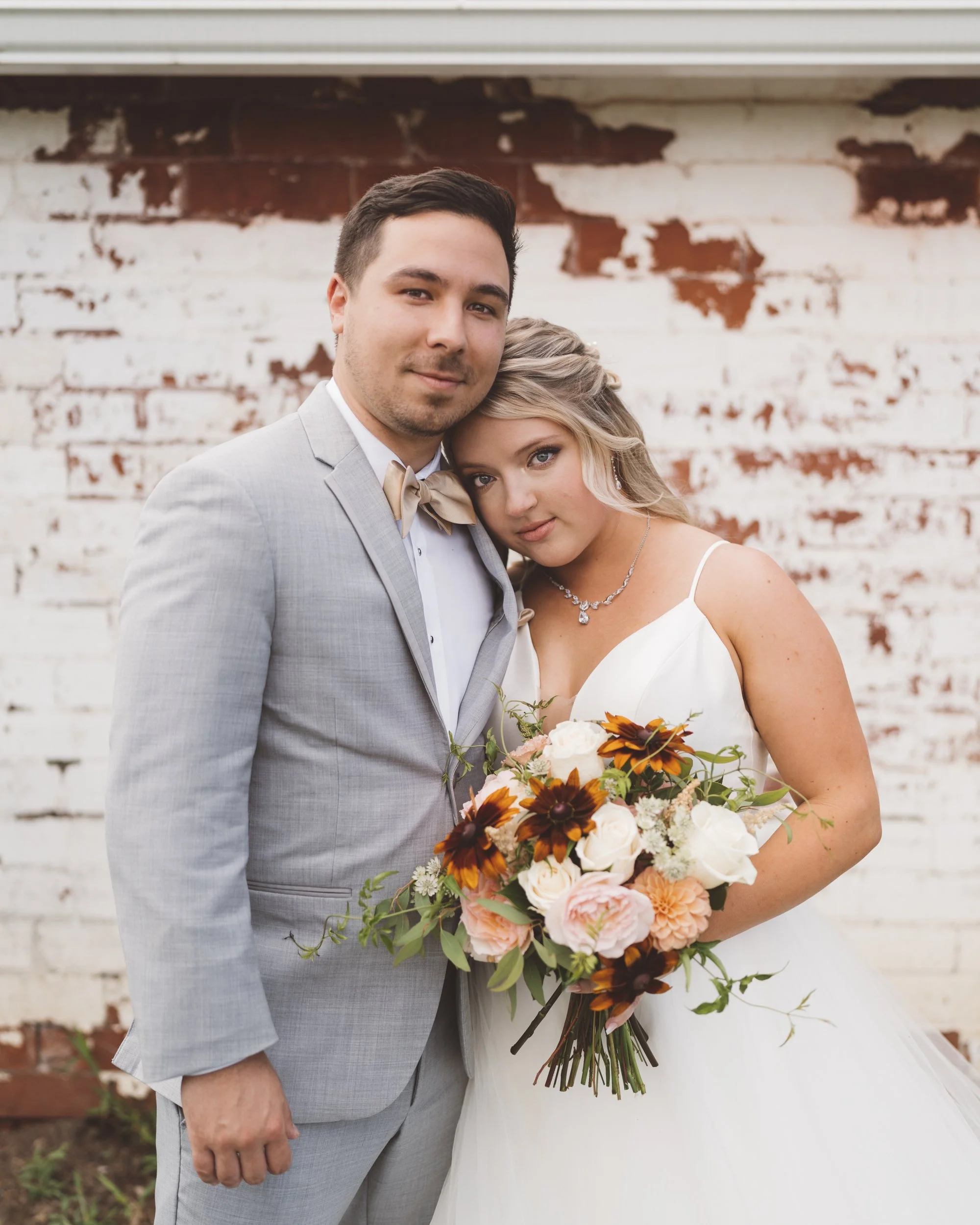 A newlywed couple standing close together, with the bride holding a large bouquet of flowers, both dressed in wedding attire in front of a weathered brick wall.