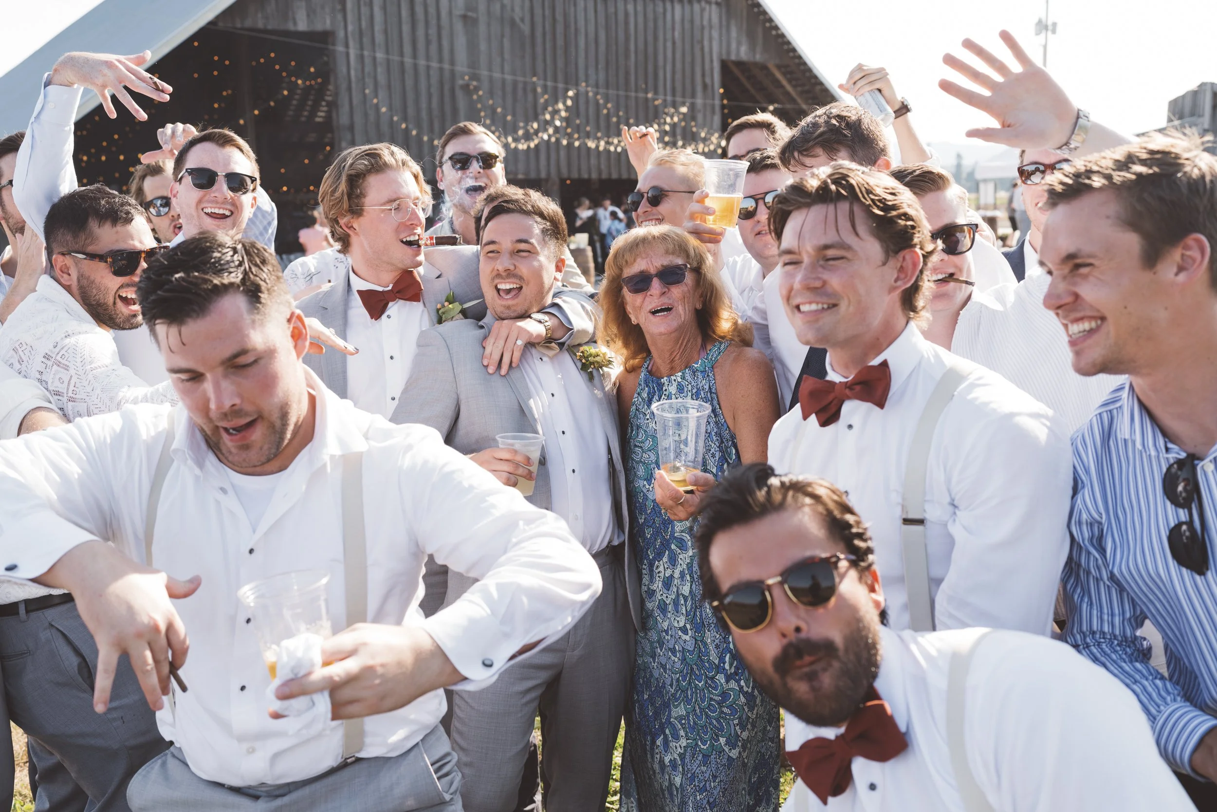 A group of people at a wedding celebration outdoors, smiling and dancing, some holding drinks, with string lights and a building in the background.