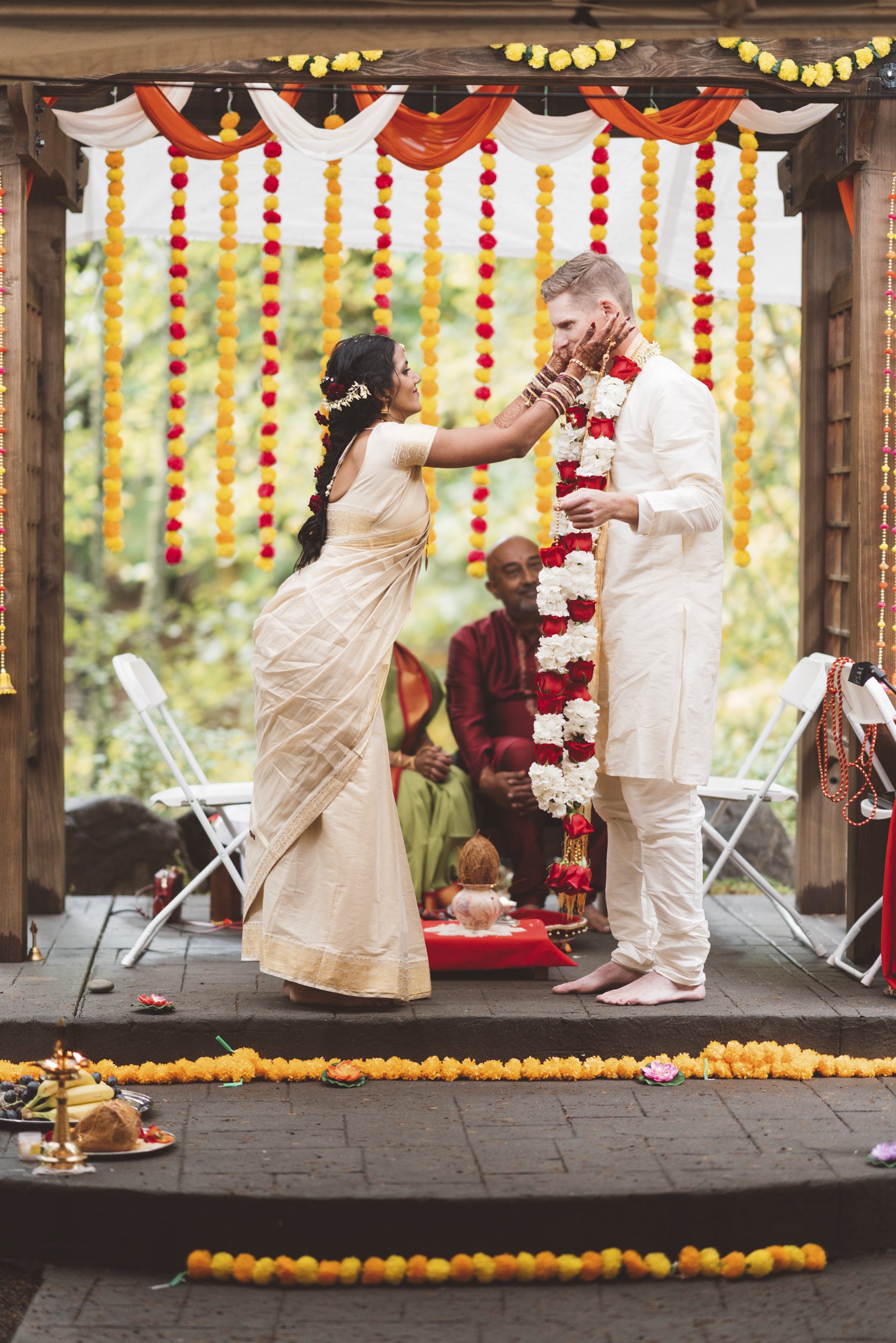A Hindu wedding ceremony with a woman in traditional attire applying a tilak on a man's forehead, both dressed in white, under decorated outdoor canopy with marigold garlands, while a priest observes in the background.