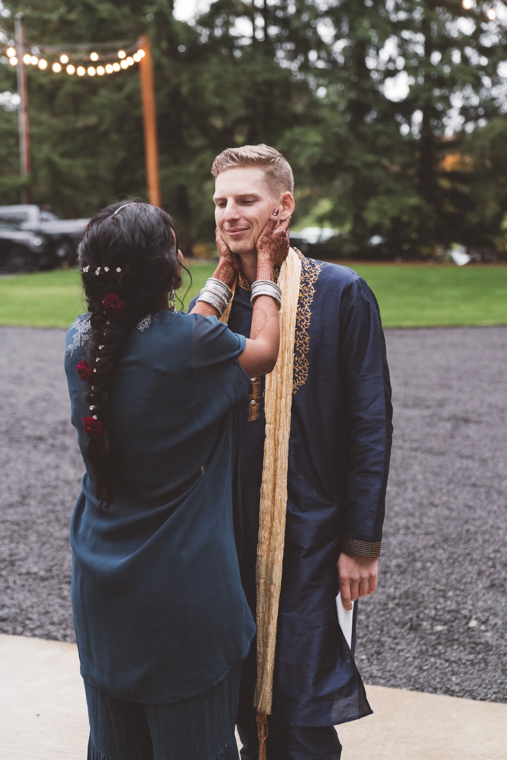 A woman with henna tattoos and jewelry gently holds a man's face, both dressed in traditional Indian attire outdoors during evening with string lights overhead, greenery in the background, and a gravel ground.