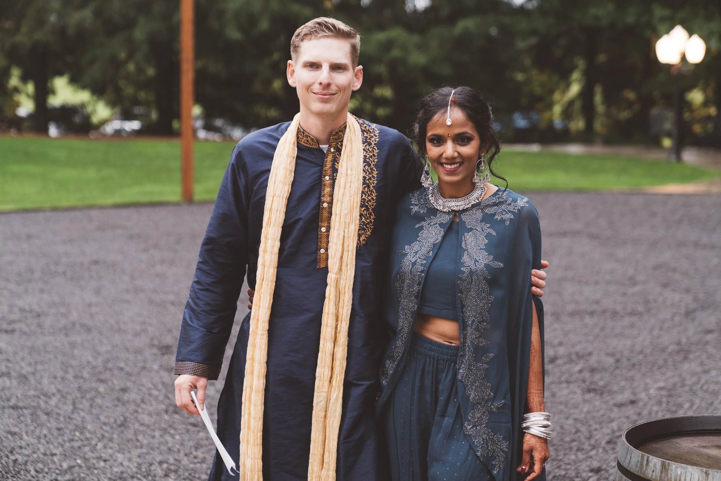 A man and woman dressed in traditional Indian attire standing outdoors on a gravel area with trees and lamp posts in the background, smiling at the camera.