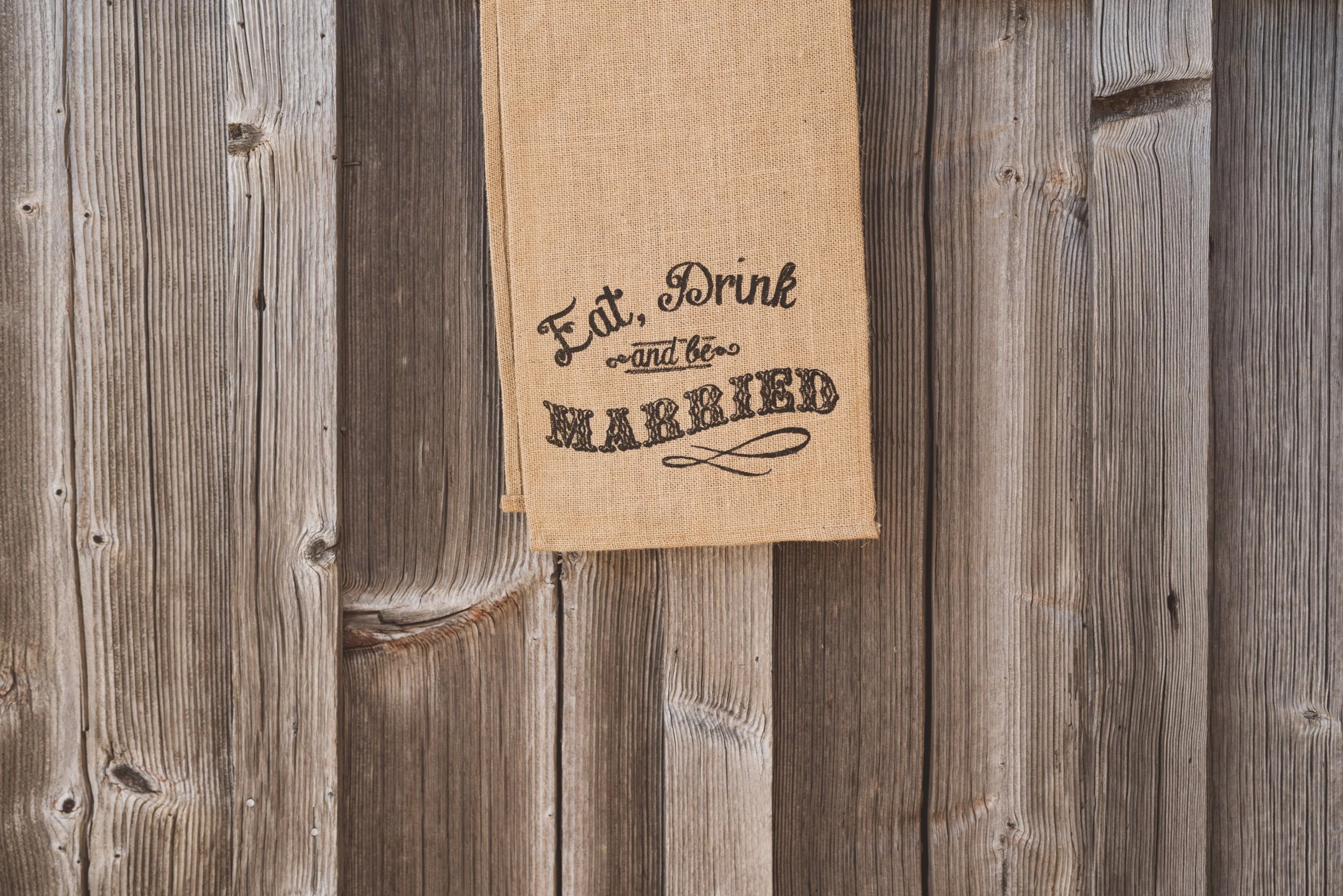 A cloth towel hanging on a rustic wooden wall with the words 'Eat, Drink and be Married' printed on it.