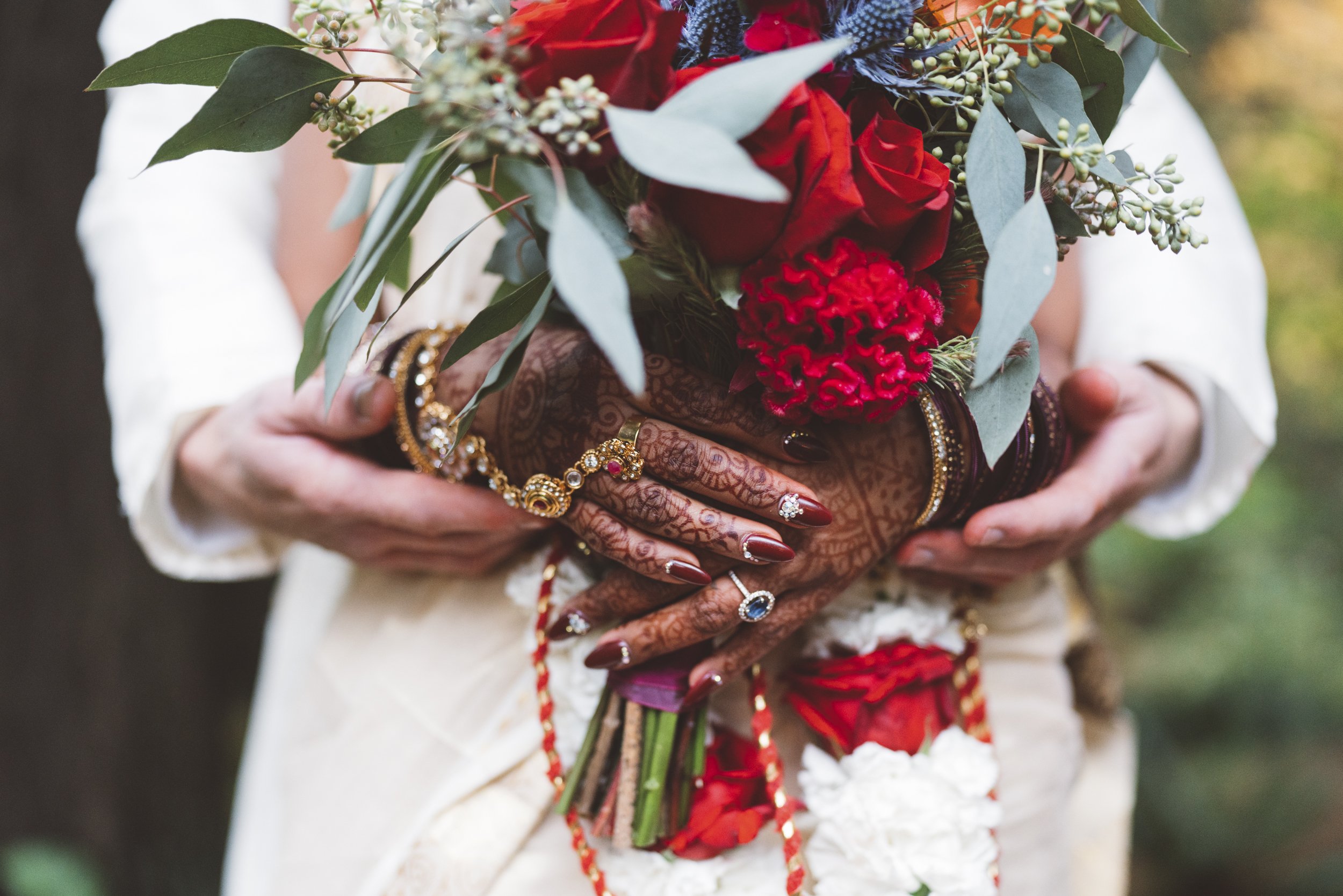 Close-up of a woman's hands holding a bouquet of red roses, carnations, and greenery, with intricate henna designs on her hands and decorated with rings and bracelets, during a traditional Indian wedding.