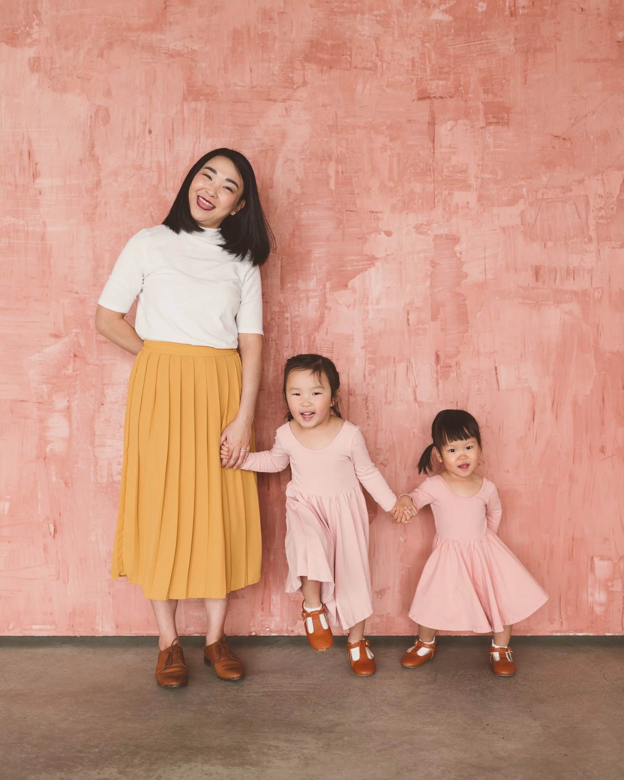 A woman and two young girls smiling and holding hands in front of a pink textured wall.