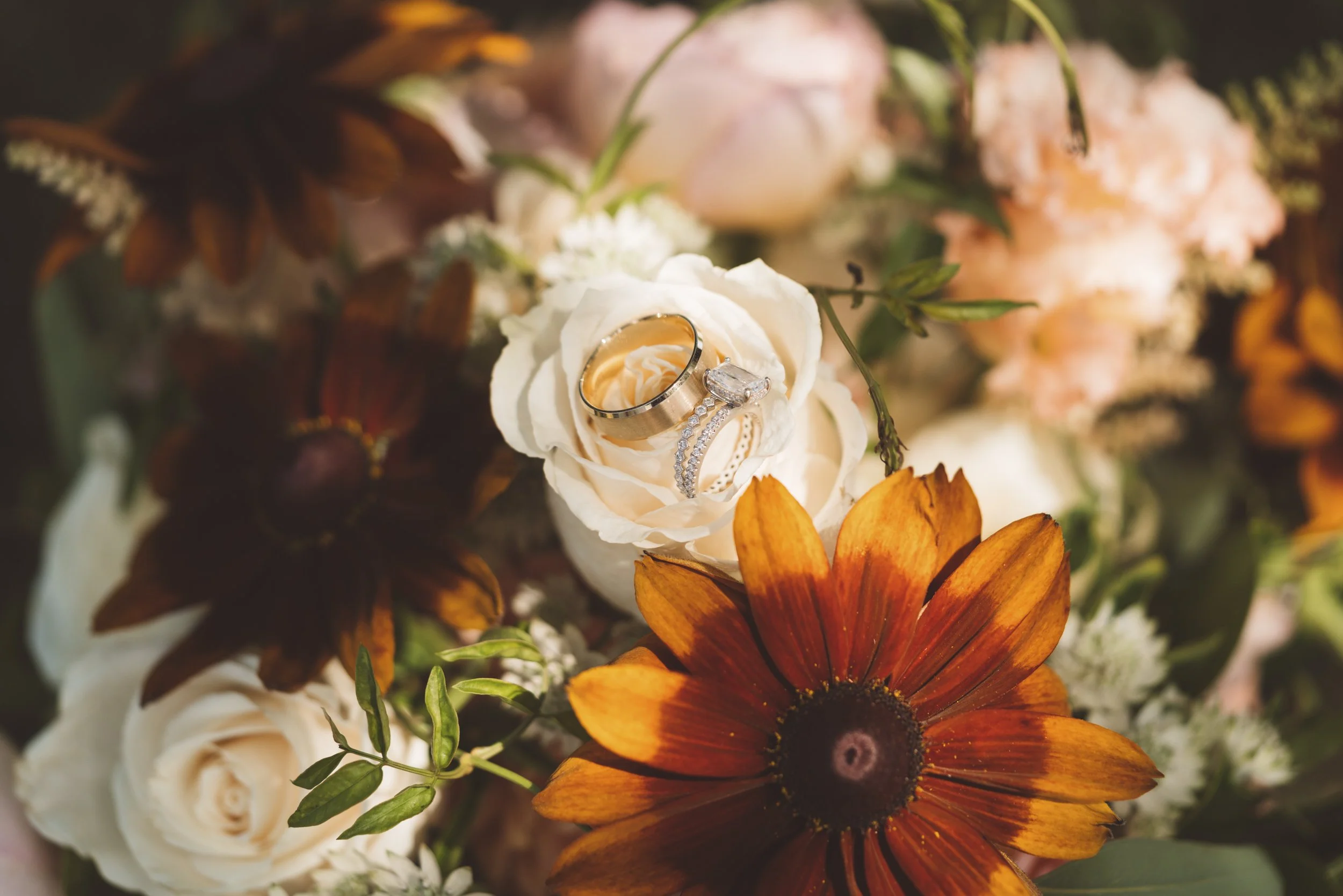 A wedding bouquet with white roses, orange and dark red sunflowers, and other flowers, featuring wedding rings resting on one of the roses.