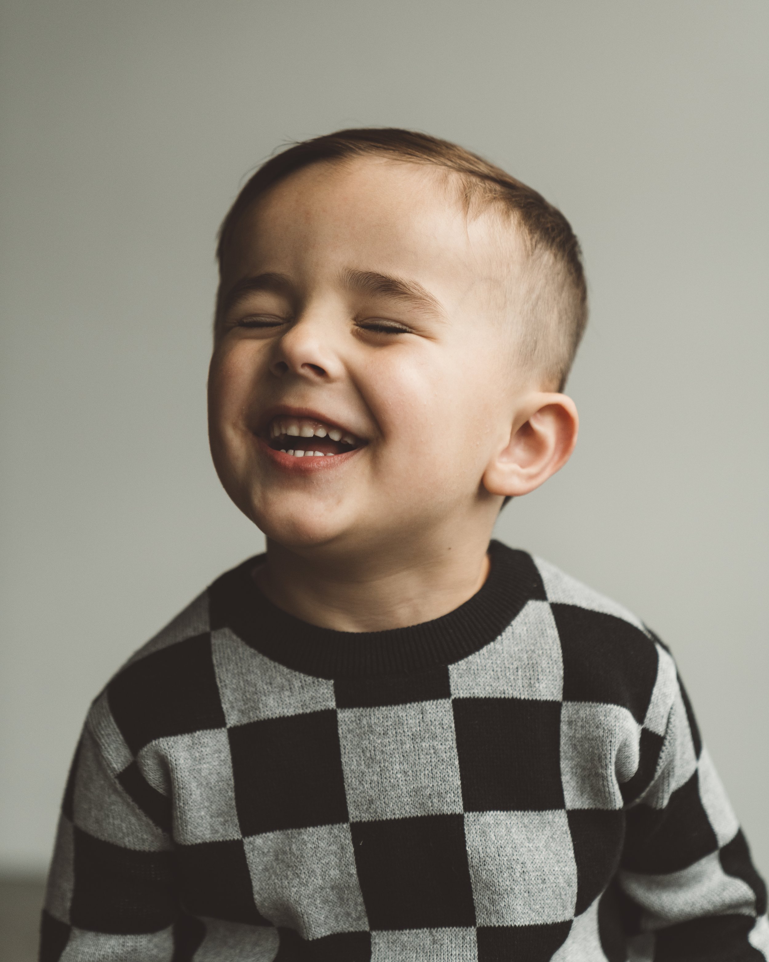 A young boy with short brown hair and a checkered black and gray sweater laughing with eyes closed and mouth open, showing teeth.