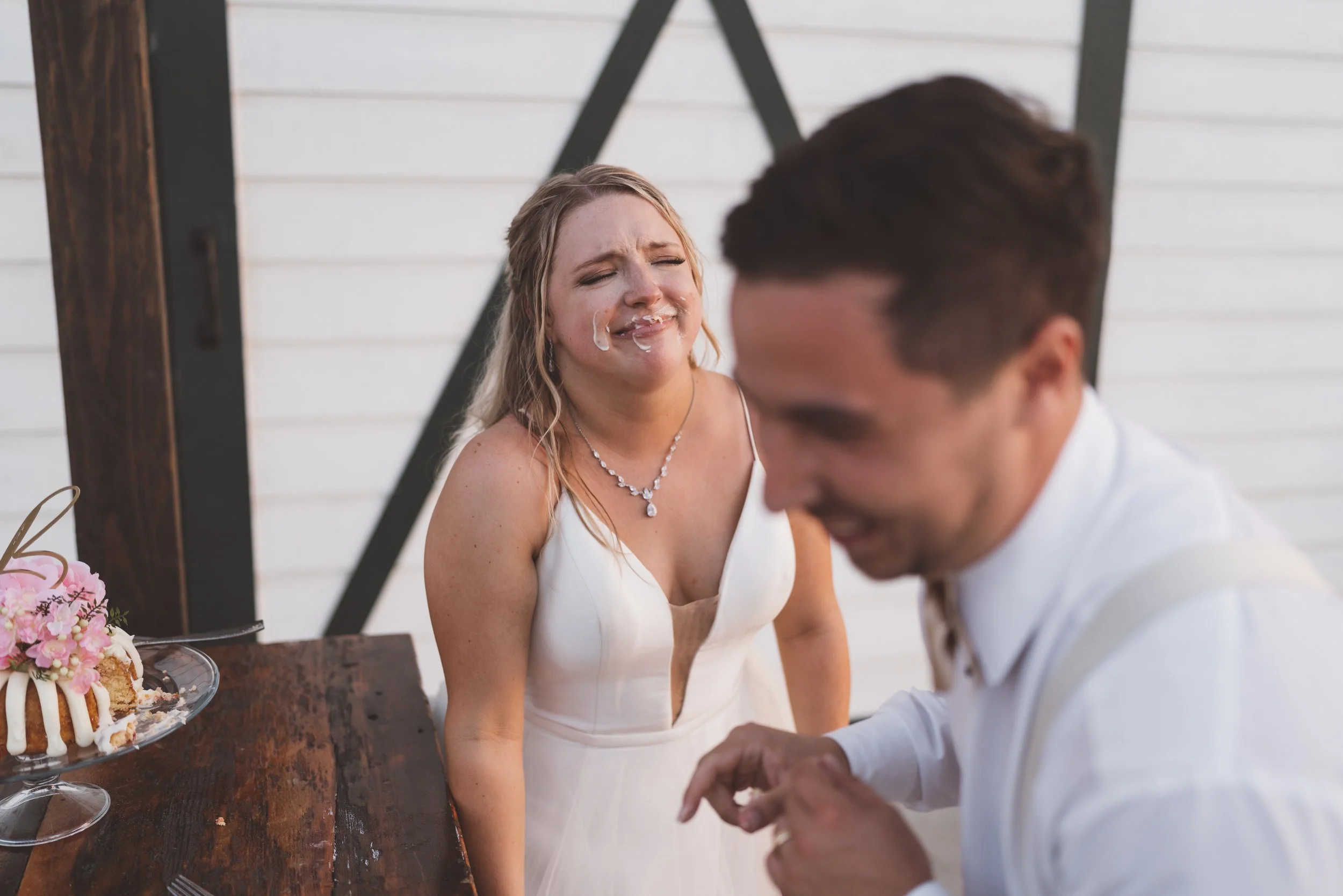 A woman in a white dress crying and smiling, with a man in white attire in front of her, at a wedding reception with a cake on a table nearby.