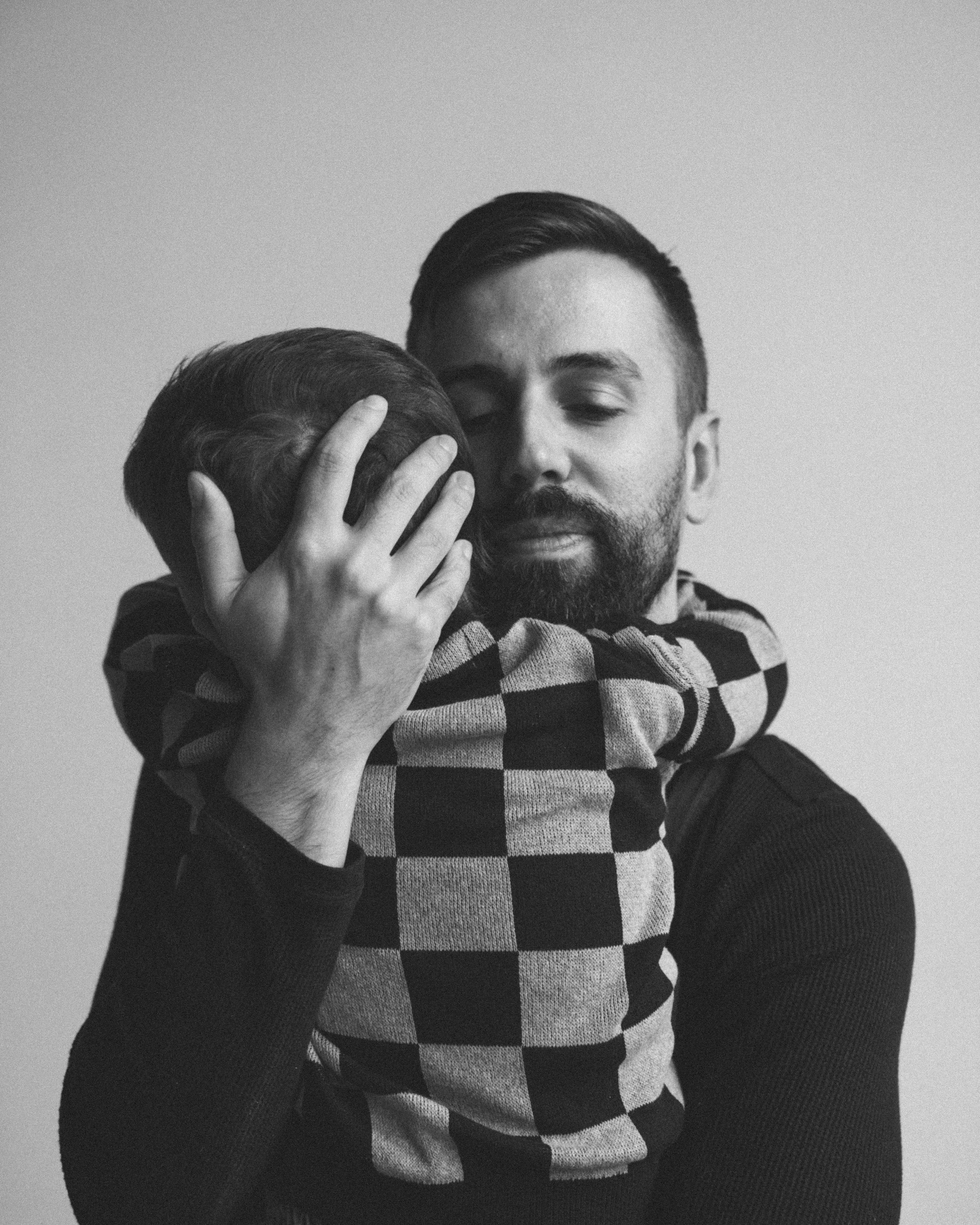 A man with a beard holds a young boy in a checkered shirt, hugging him tightly with his eyes closed.