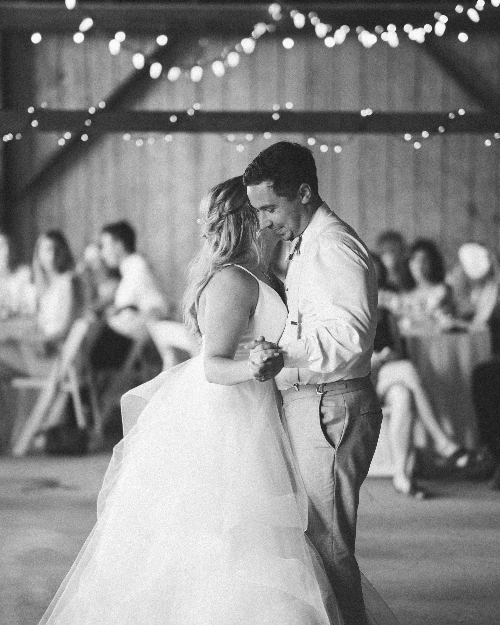 A black-and-white photo of a couple dancing at a wedding reception, with string lights overhead and guests in the background.