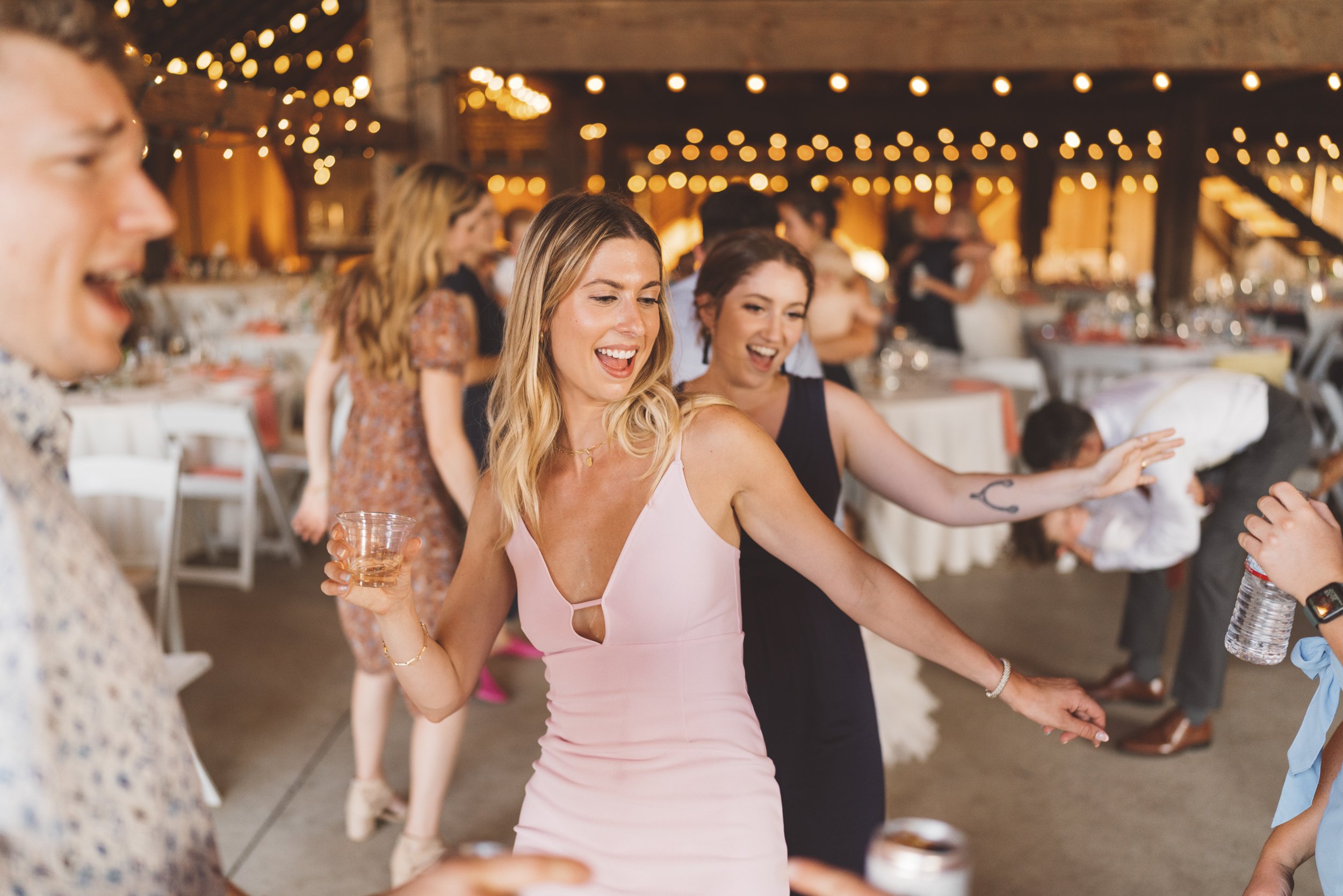 Women dancing and having fun at a party or celebration in a decorated venue with string lights and tables in the background.
