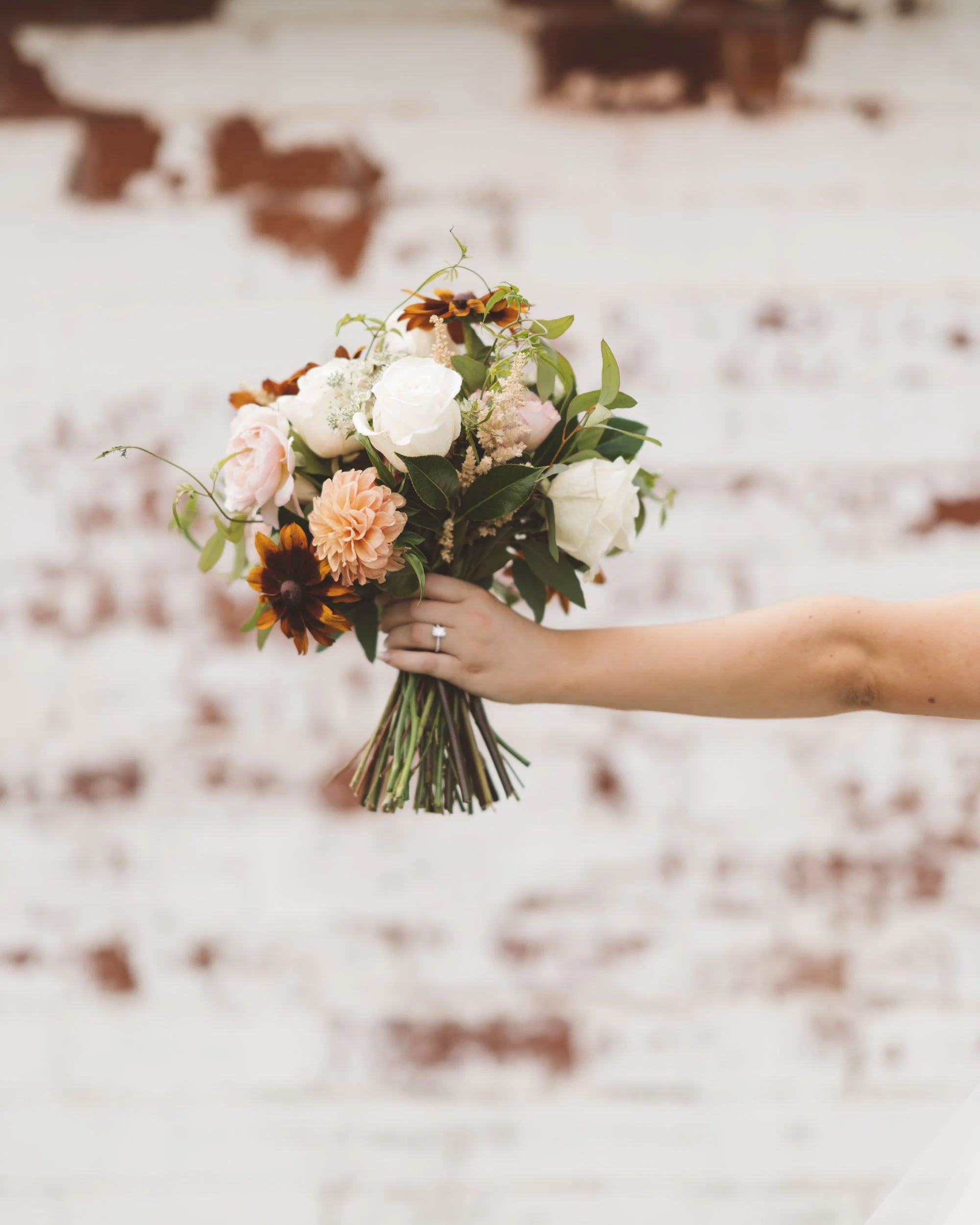Person holding a bouquet of mixed flowers including white roses, pink peonies, orange dahlias, and brown-black flowers with green foliage, against a white brick wall background.