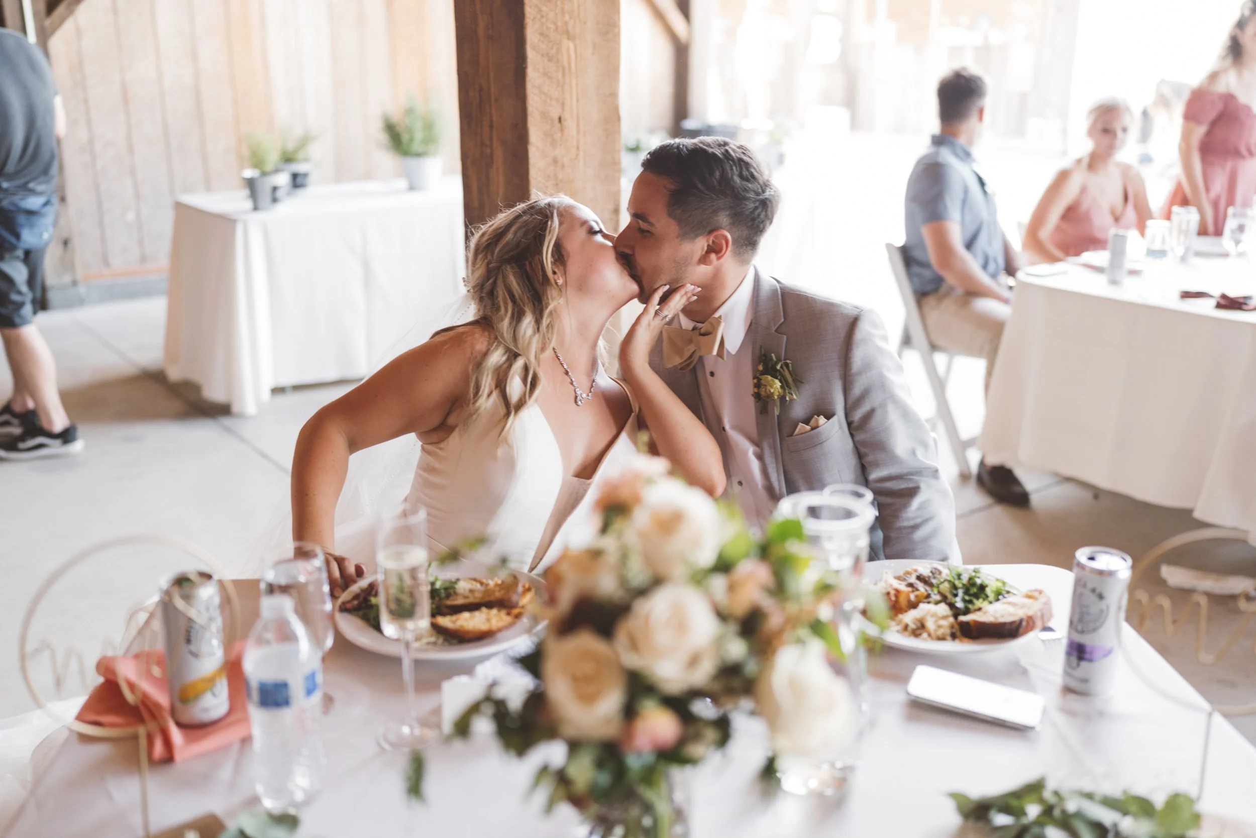 A bride and groom share a kiss at their wedding reception, seated at a decorated table with flowers, food, and drinks around them.
