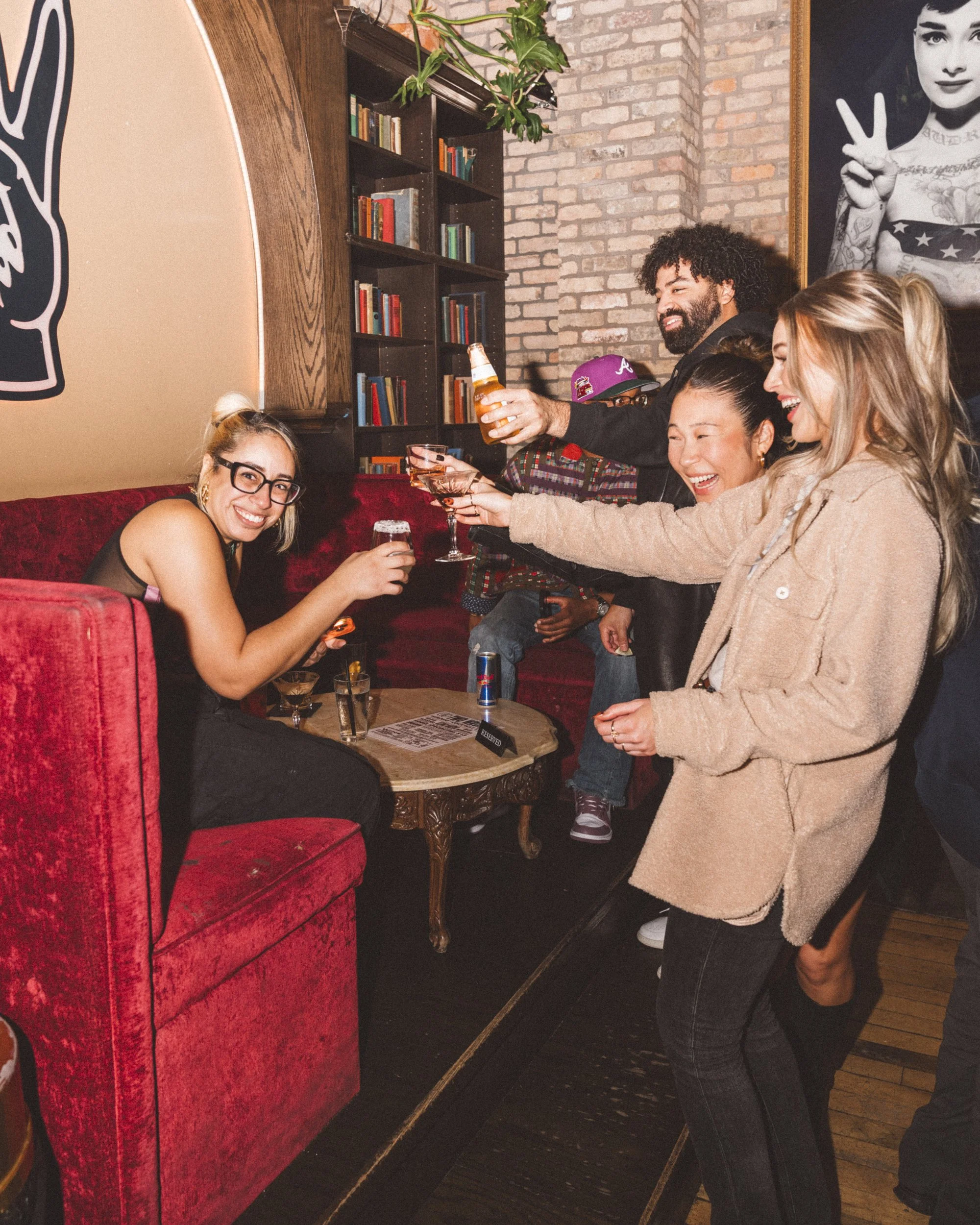 Group of friends celebrating and toasting drinks in a cozy bar with bookshelves and artwork in the background.