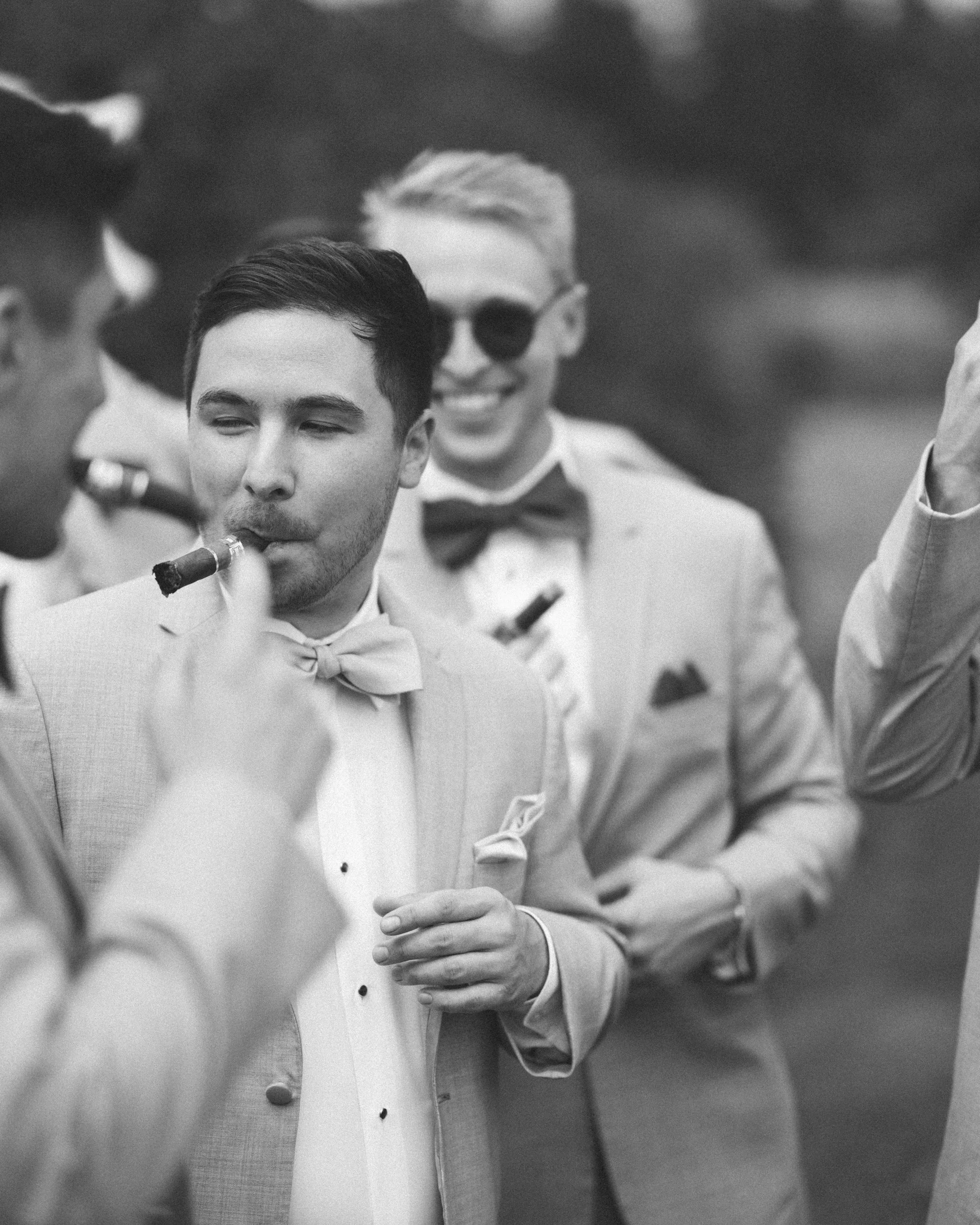 Black and white photo of a group of men in tuxedos enjoying cigars at an outdoor event.