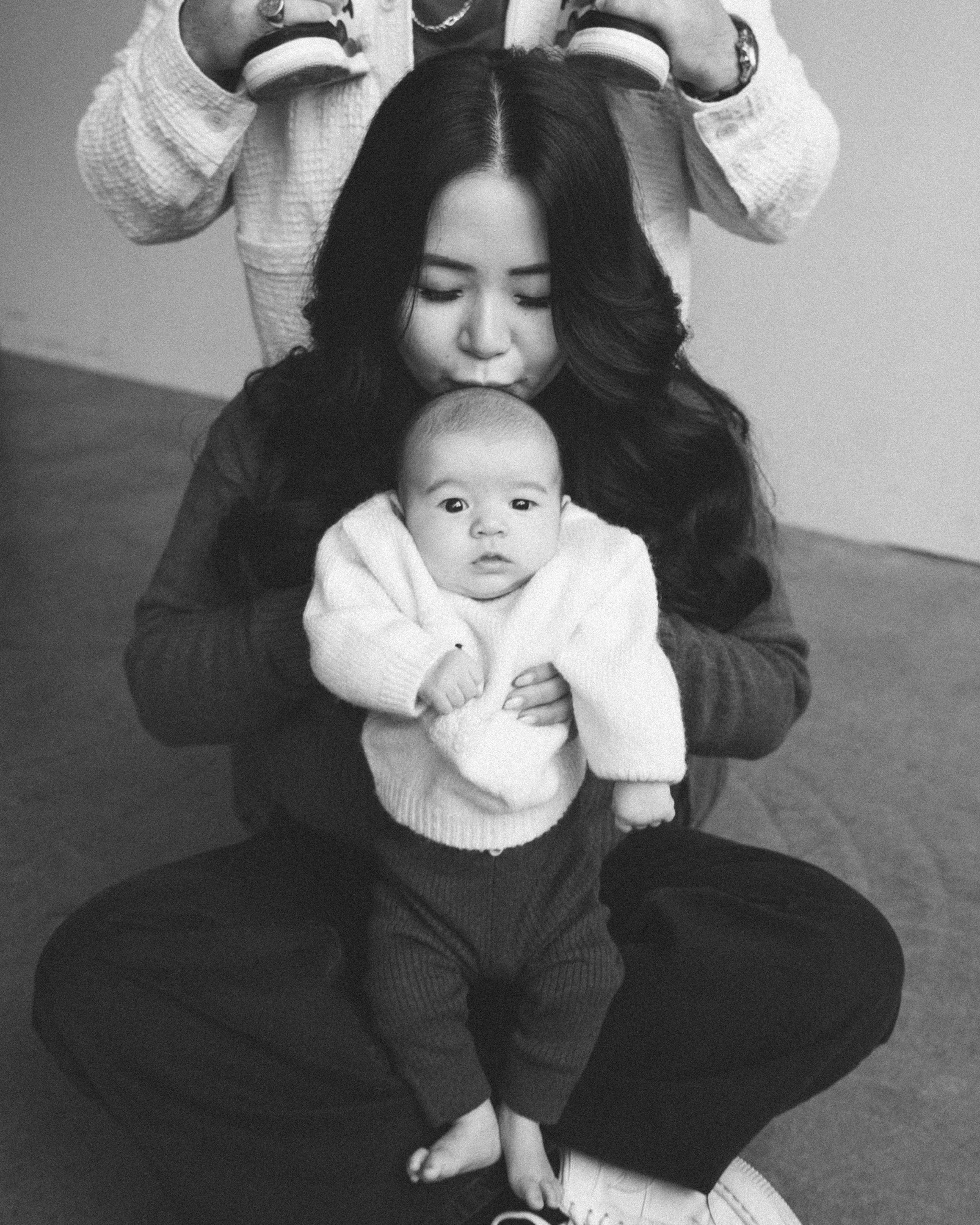 A woman holding a baby in her lap, with another person standing behind them, partially visible. The woman is kissing the baby's head, and the baby looks directly at the camera. The scene is in black and white.
