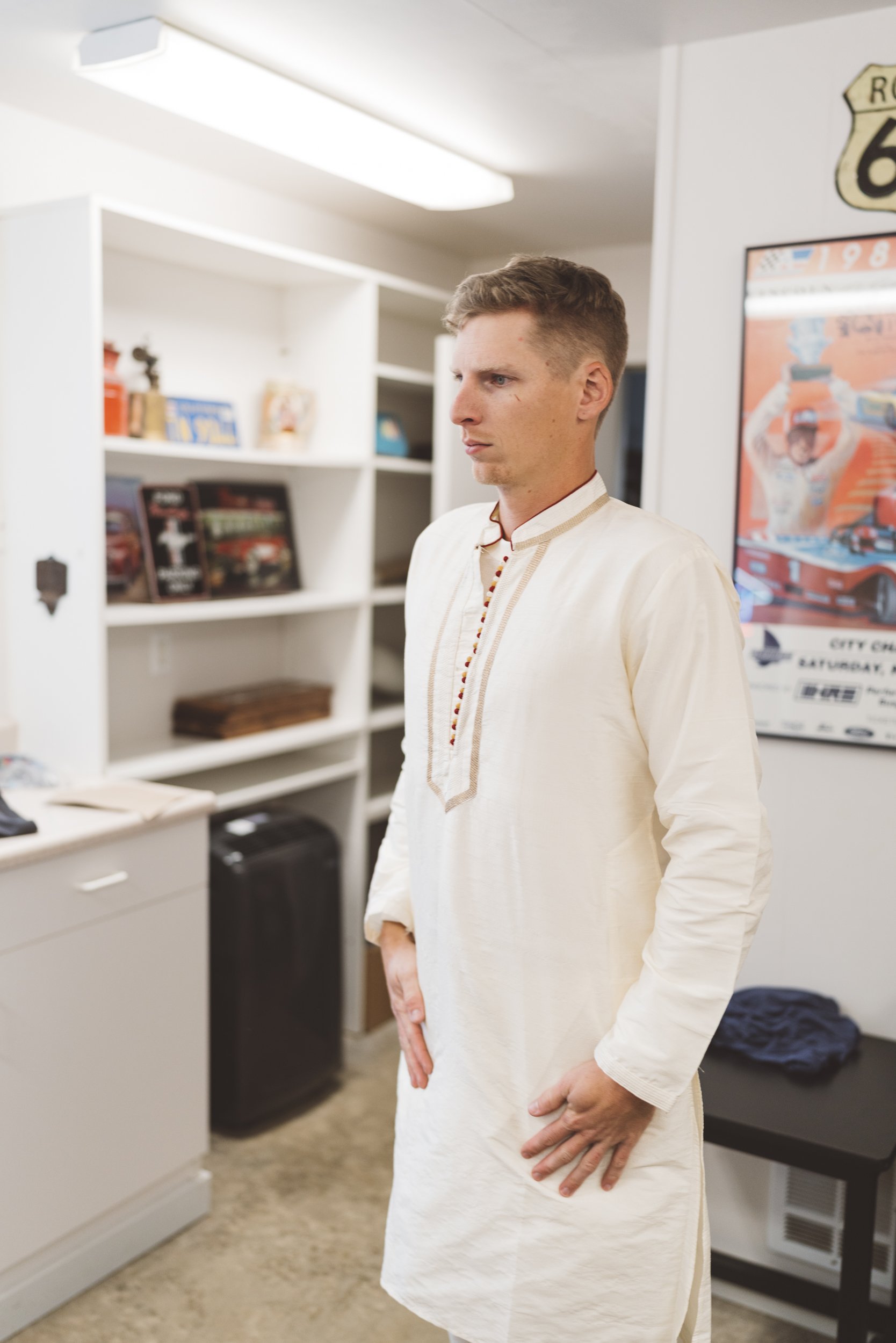 Man wearing traditional Indian kurta, standing in a room with white shelves and a poster of race cars on the wall.