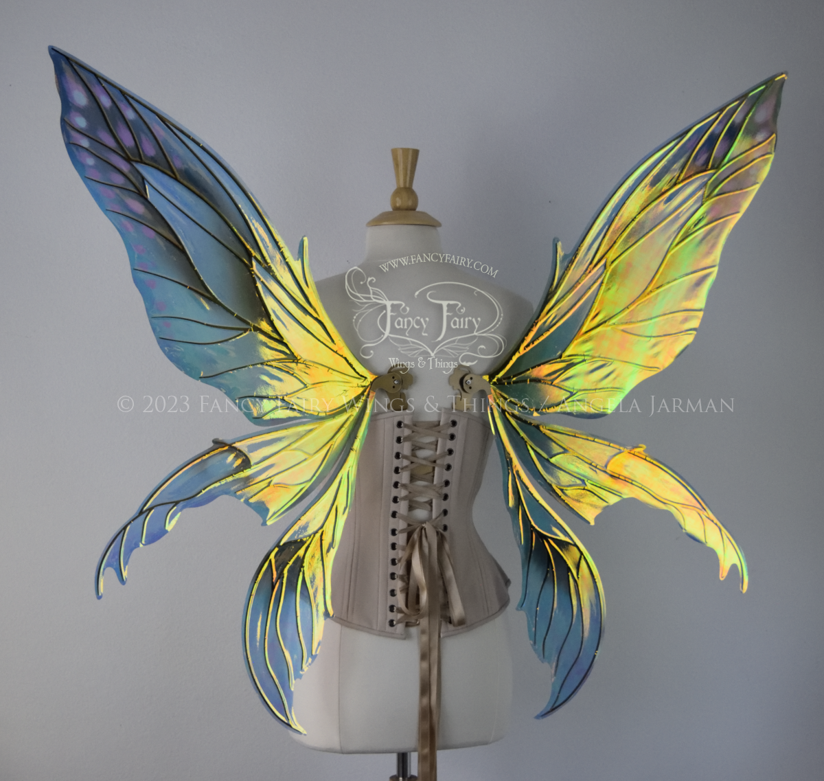  Back view of large fairy wings in iridescent green and blue with pink spots near the tips of the upper panels, with gold veins, worn on a dressform 