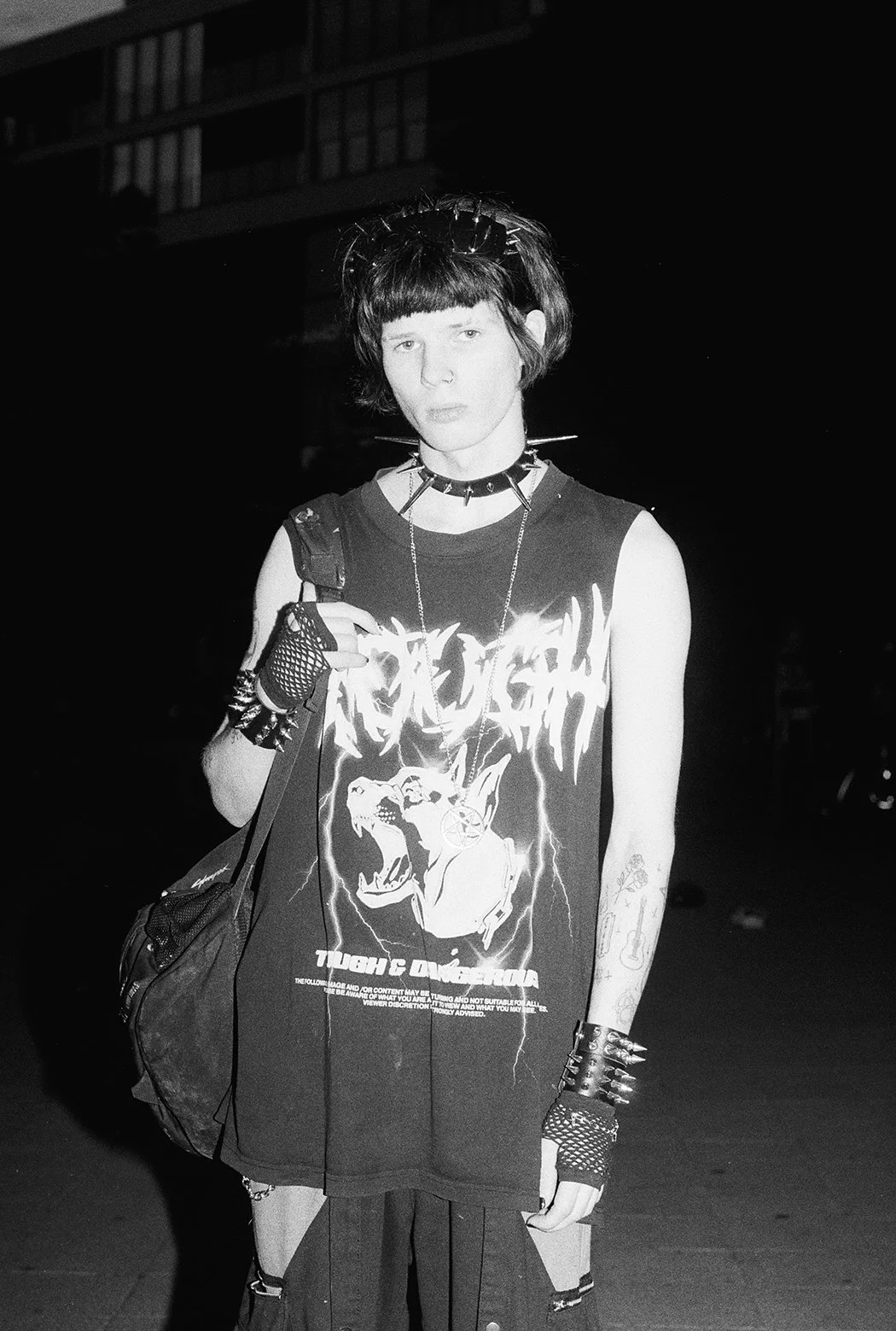 A shirtless young man with curly hair and a beard standing on a city street, wearing headphones around his neck, with a tattoo of Jesus on the cross on his chest, and holding a checkered shirt.