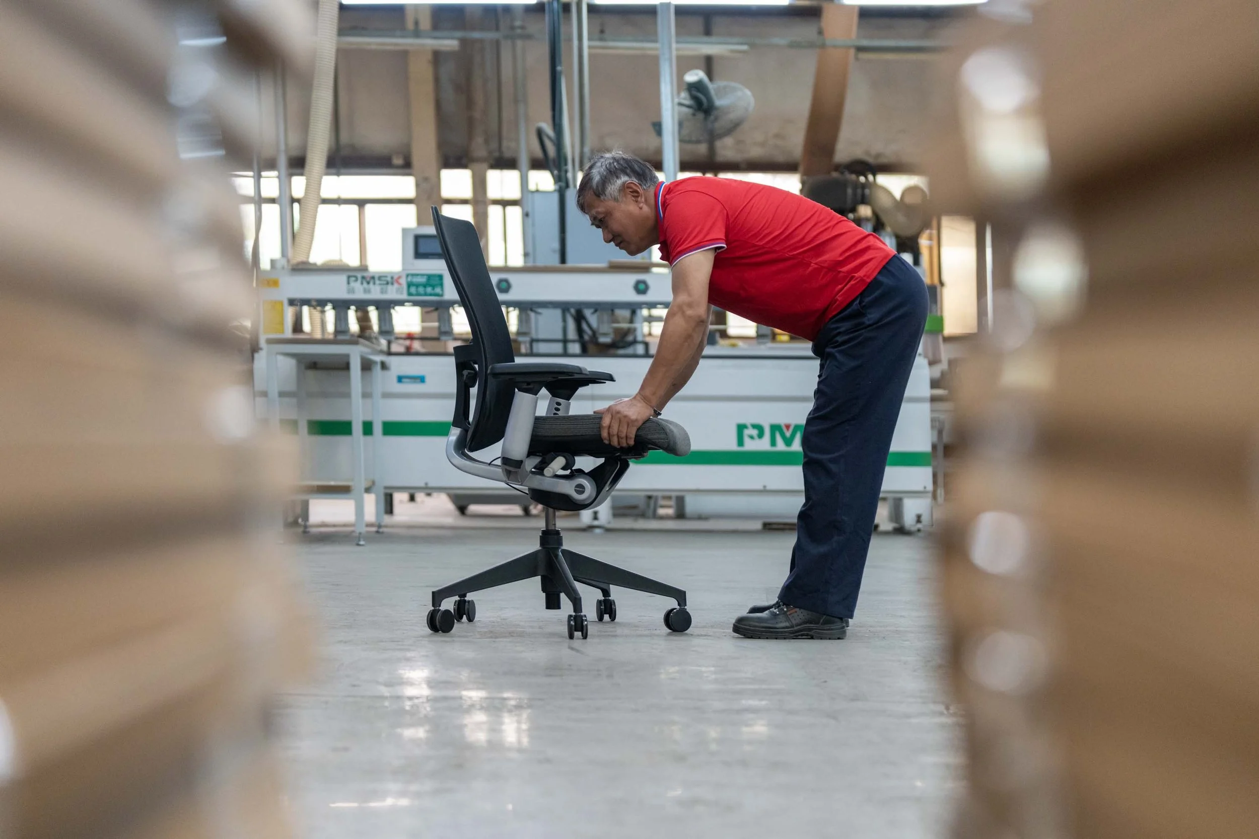 A man in a red shirt and dark pants bending over to adjust a black office chair in a spacious industrial or factory setting.
