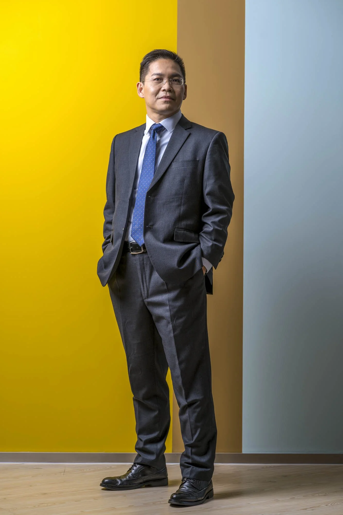 A man wearing a dark suit, light blue shirt, and blue tie stands with his hands in his pockets against a colorful background of yellow, brown, and blue walls, on a wooden floor.