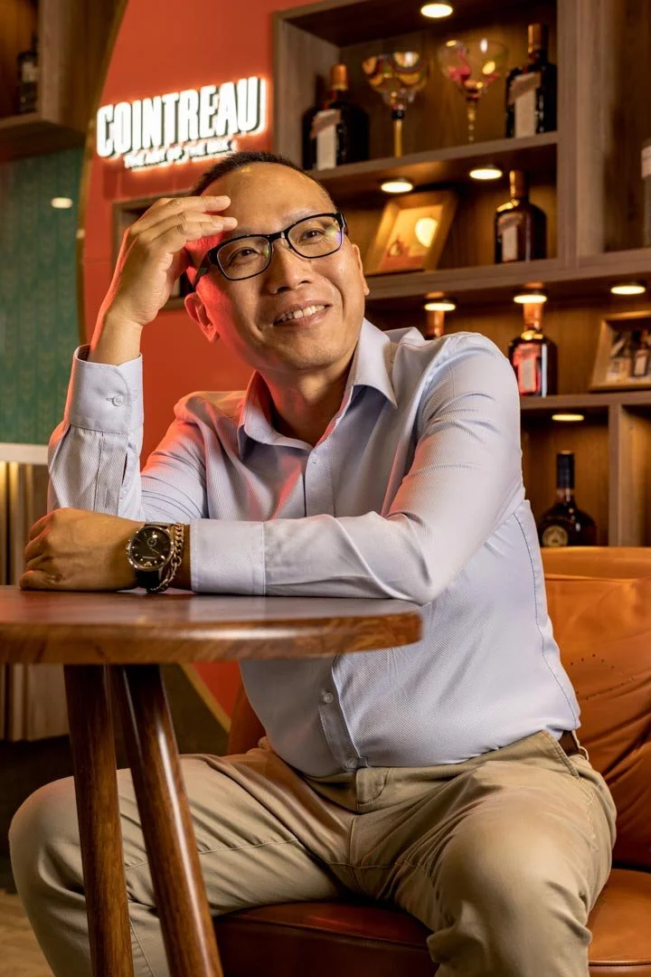 A man wearing glasses, a light-colored dress shirt, and khaki pants sitting at a wooden table in a bar or restaurant with a relaxed and smiling demeanor, with shelves of bottles and a neon sign reading 'COUNTEAU' in the background.