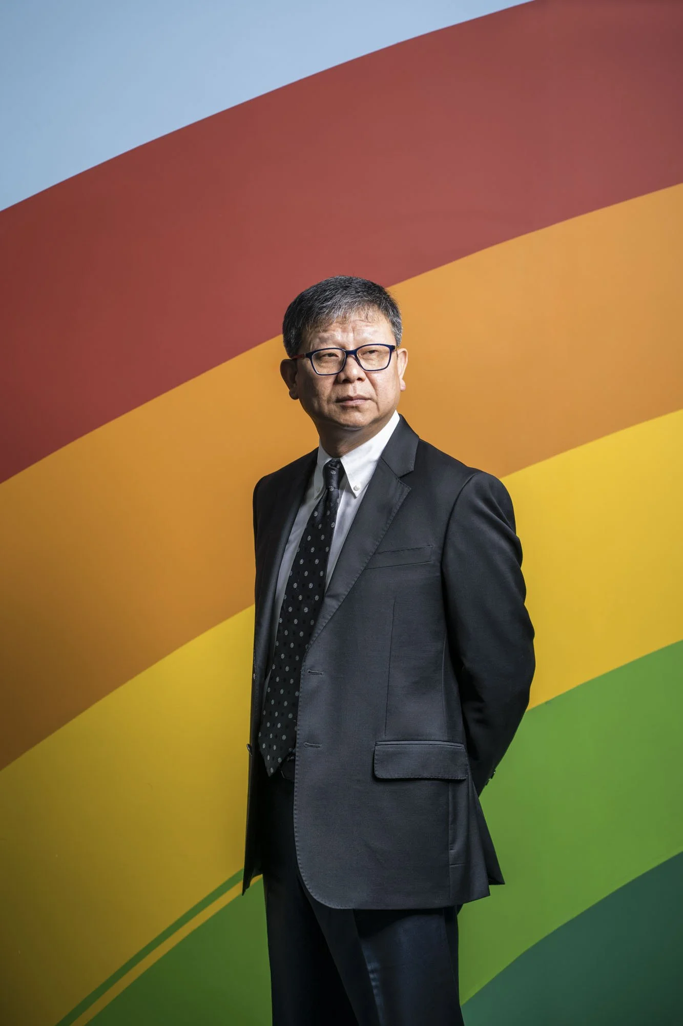 A man in a suit and tie with glasses standing in front of a colorful rainbow backdrop.